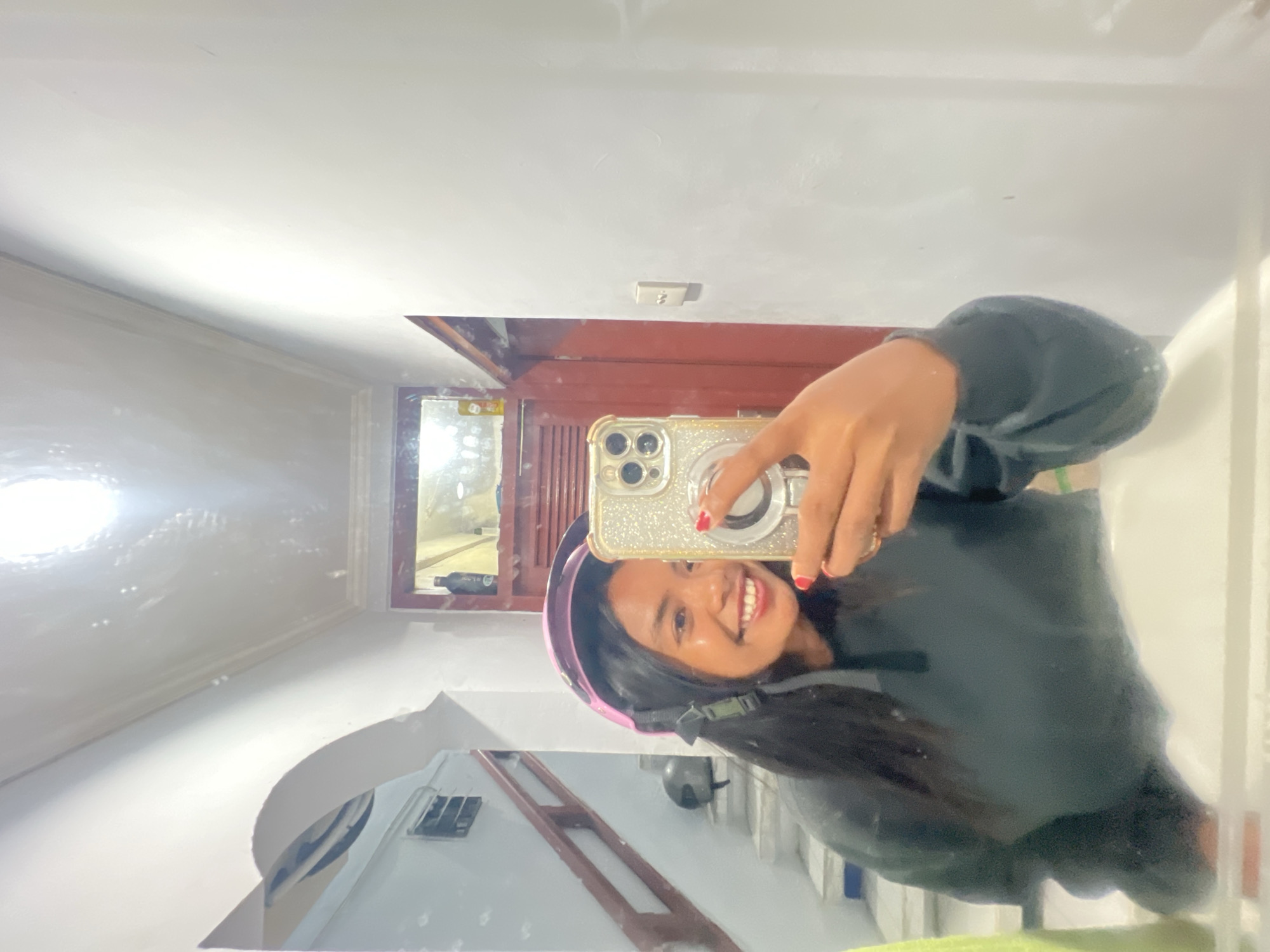 A young woman with long, dark hair is reflected in a mirror, wearing a pink helmet and a dark hoodie. She is holding a smartphone with a glittery case, taking a selfie. Her face is lit by overhead lights, revealing a wide smile and red nail polish. Behind her, in the reflection, a staircase with wooden railings is visible, along with a helmet on a step. The room appears to be a bathroom or hallway, with white walls, an arched doorway, and a red door. The overall impression is of a casual, indoor setting.
