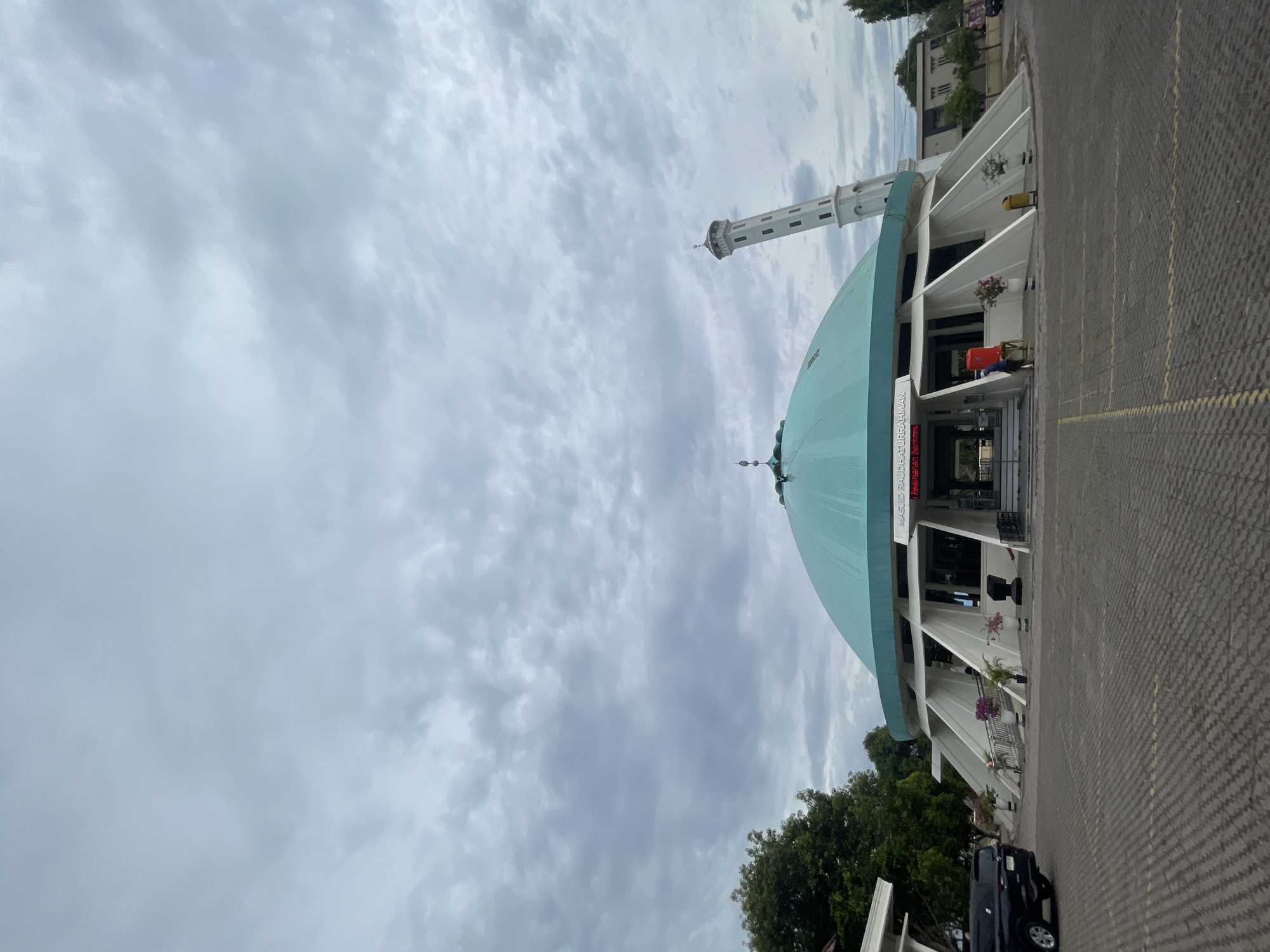The image shows the exterior of a mosque with a large, teal-colored dome and a tall, white minaret. The mosque is situated in a paved courtyard. A black SUV is parked to the left of the mosque. A person in a red shirt is visible near the entrance of the mosque. The sky is overcast with gray clouds. The text on the mosque reads "MASJID RAUDHATURRAHMAN" and "Senyuman Bersama".