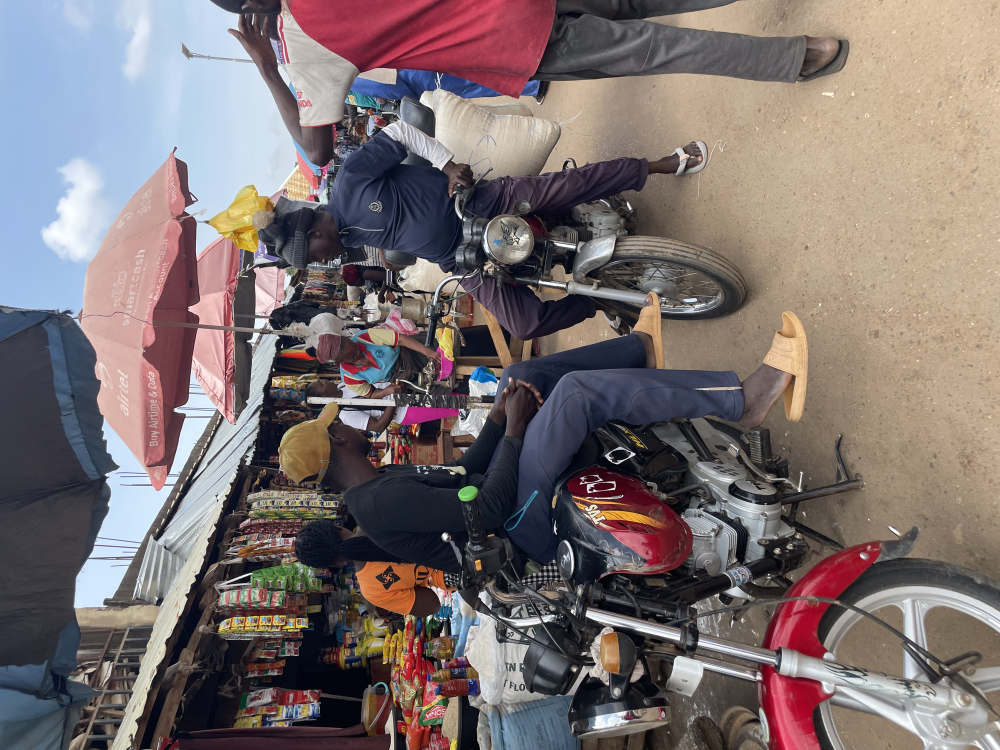 In Akwanga, Nigeria, two men are seen on motorcycles in a marketplace. One man, wearing a yellow cap and dark clothing, sits on a red and black motorcycle. The second man, in a navy blue long-sleeved shirt, sits on a silver motorcycle. They appear to be conversing. Several market stalls with various goods are visible in the background, along with red and pink umbrellas overhead. The ground is a dirt surface.