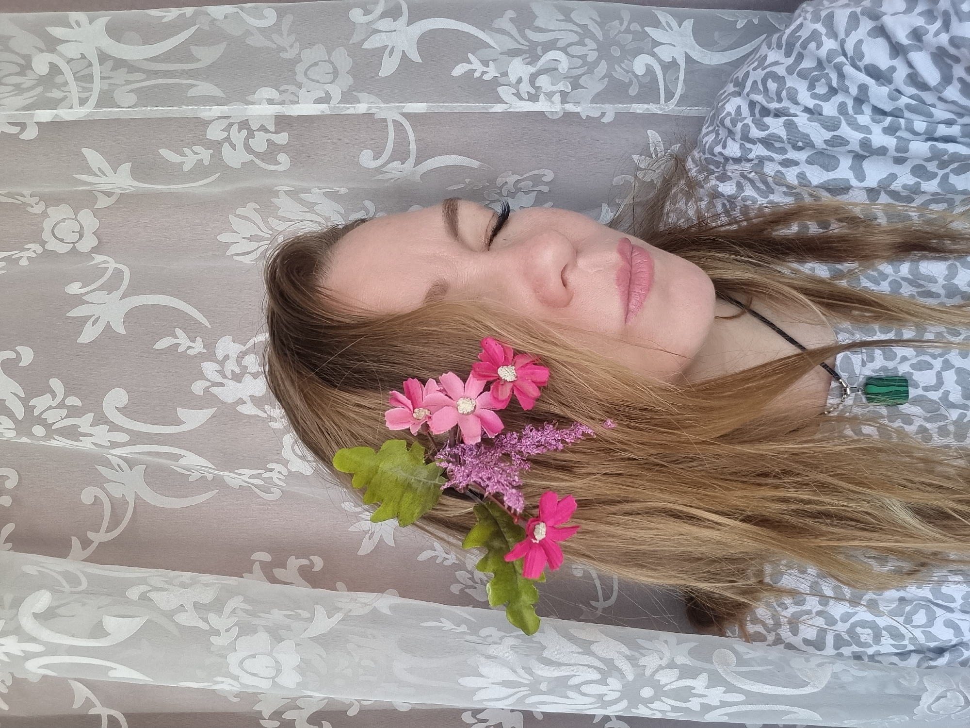 A woman is lying down. She has blonde hair with pink and purple flowers in her hair. She is wearing a necklace with a green stone pendant. She is wearing a grey and white patterned shirt. She is lying on a white floral patterned fabric.
I cannot verify the location as Chaikovsky, Russia, based on the given image.