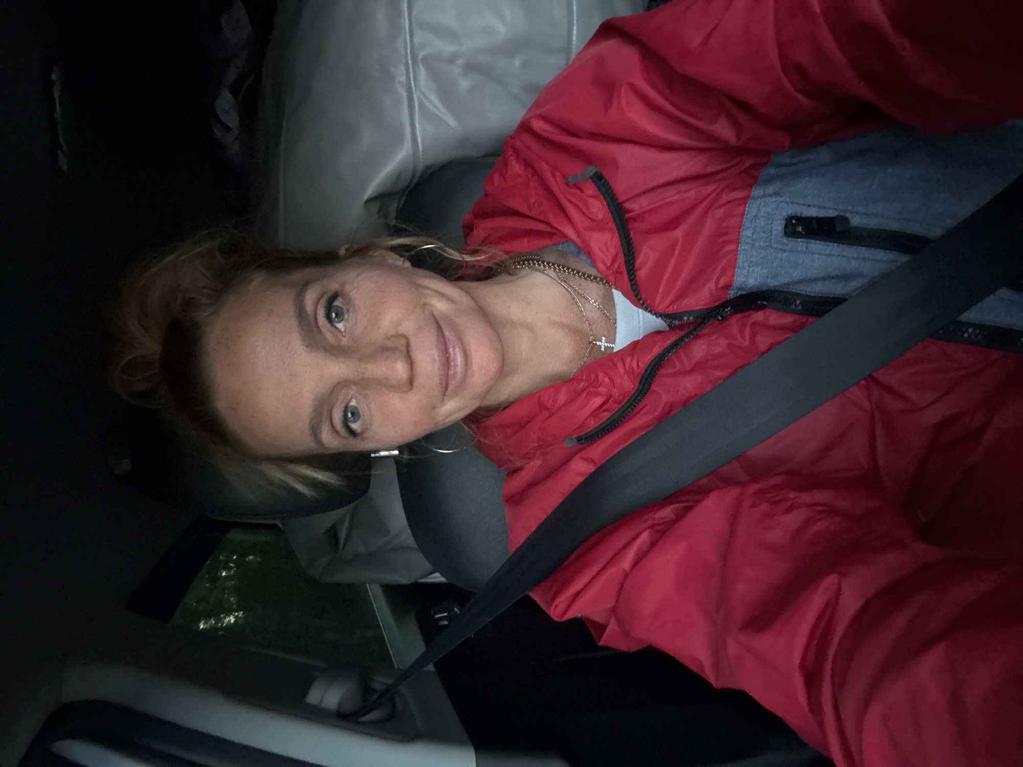 A blonde woman with freckles and her hair pulled up in a messy bun is sitting in the driver's seat of a car. She is wearing a red and grey jacket, a light blue shirt, and a necklace with a cross pendant. Her left arm is visible in the foreground, with a seatbelt across her chest. The interior of the car is visible, including the headrest of the passenger seat and some luggage in the back. The window on the left shows a dark, wooded area, suggesting a rural or suburban location, possibly on the outskirts of Ларионово, Russia, given the mention. The lighting suggests it is either dawn or dusk.