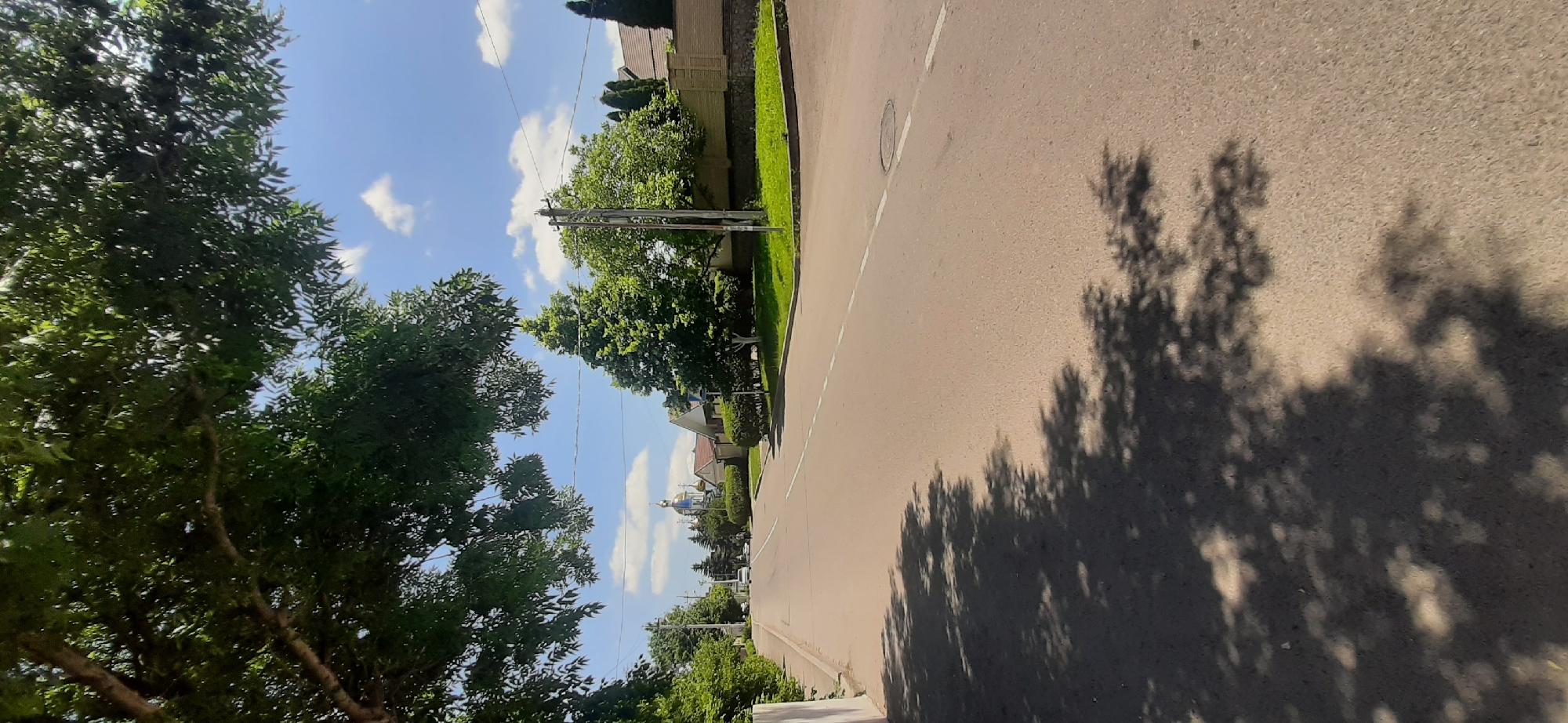The image depicts a street scene in Yessentukskaya, Russia, on a bright, sunny day. A paved road runs through the center of the frame, flanked by green trees and manicured lawns. In the distance, a church with a blue dome and golden cross is visible. Several cars are parked along the roadside, and a few pedestrians can be seen walking. The sky is a clear blue, dotted with fluffy white clouds. The overall impression is one of a peaceful, rural setting on a pleasant summer day.