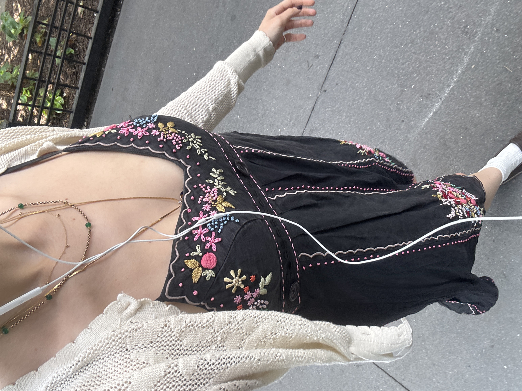 A person is seen from the chest down, wearing a black dress with floral embroidery, a cream-colored cardigan, white socks, and brown shoes. The person is standing on a paved sidewalk in an urban environment, likely New York City, as indicated by the presence of a black metal fence with greenery behind it, characteristic of city park railings. The person is wearing earbuds connected to a device not visible in the frame. The image is taken from a slightly elevated angle, looking down at the subject.