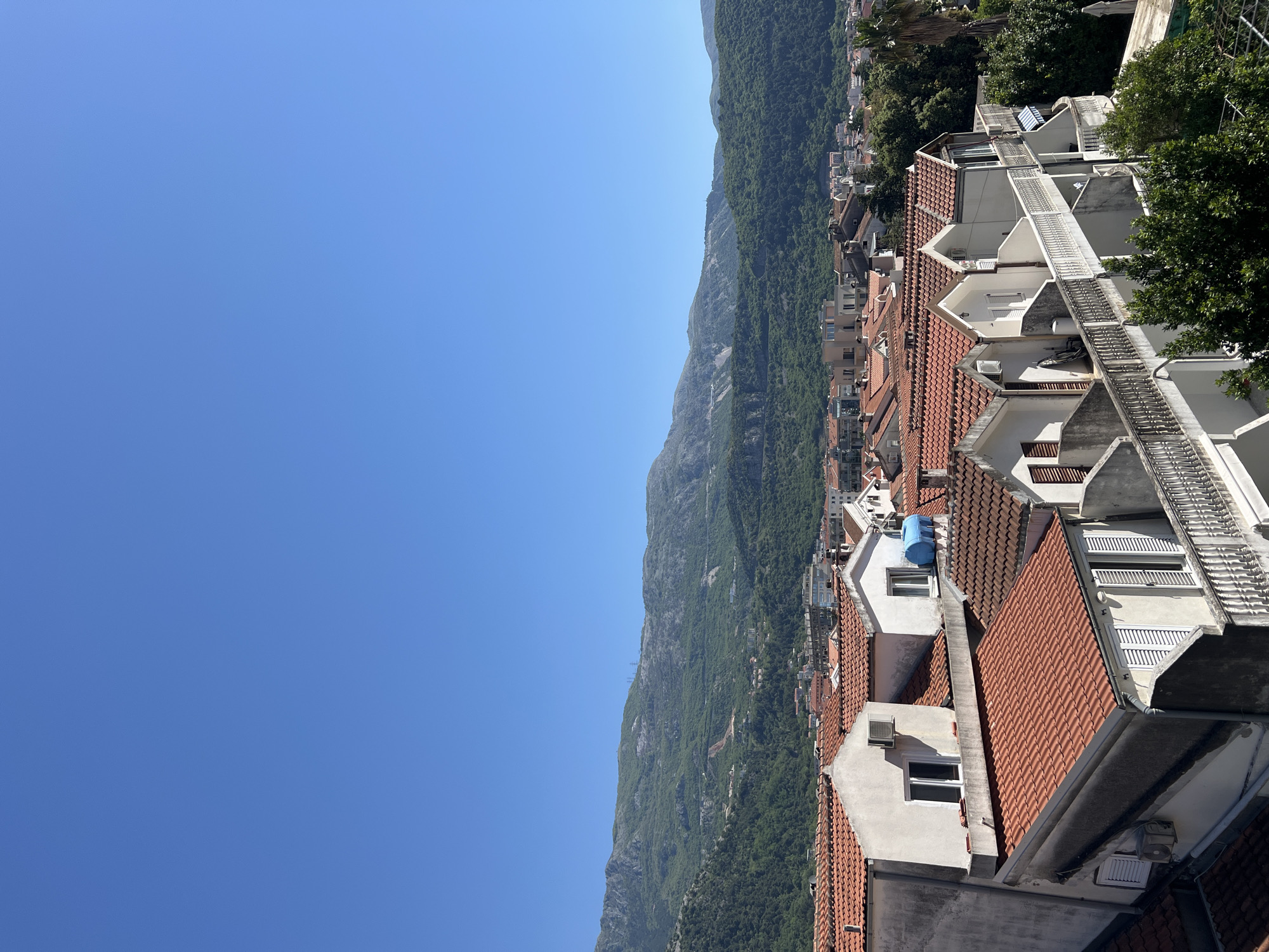 The image shows an elevated view of Budva, Montenegro, focusing on a cluster of buildings with red-tiled roofs. These buildings are situated on a hillside, leading down towards a mountain range covered in dense green vegetation. The sky above is a clear, bright blue. The architecture is predominantly white with red roof tiles. Visible details include windows, staircases, and some modern amenities, such as air conditioning units, installed on the buildings. The overall impression is that of a densely built residential area in a mountainous coastal setting.
