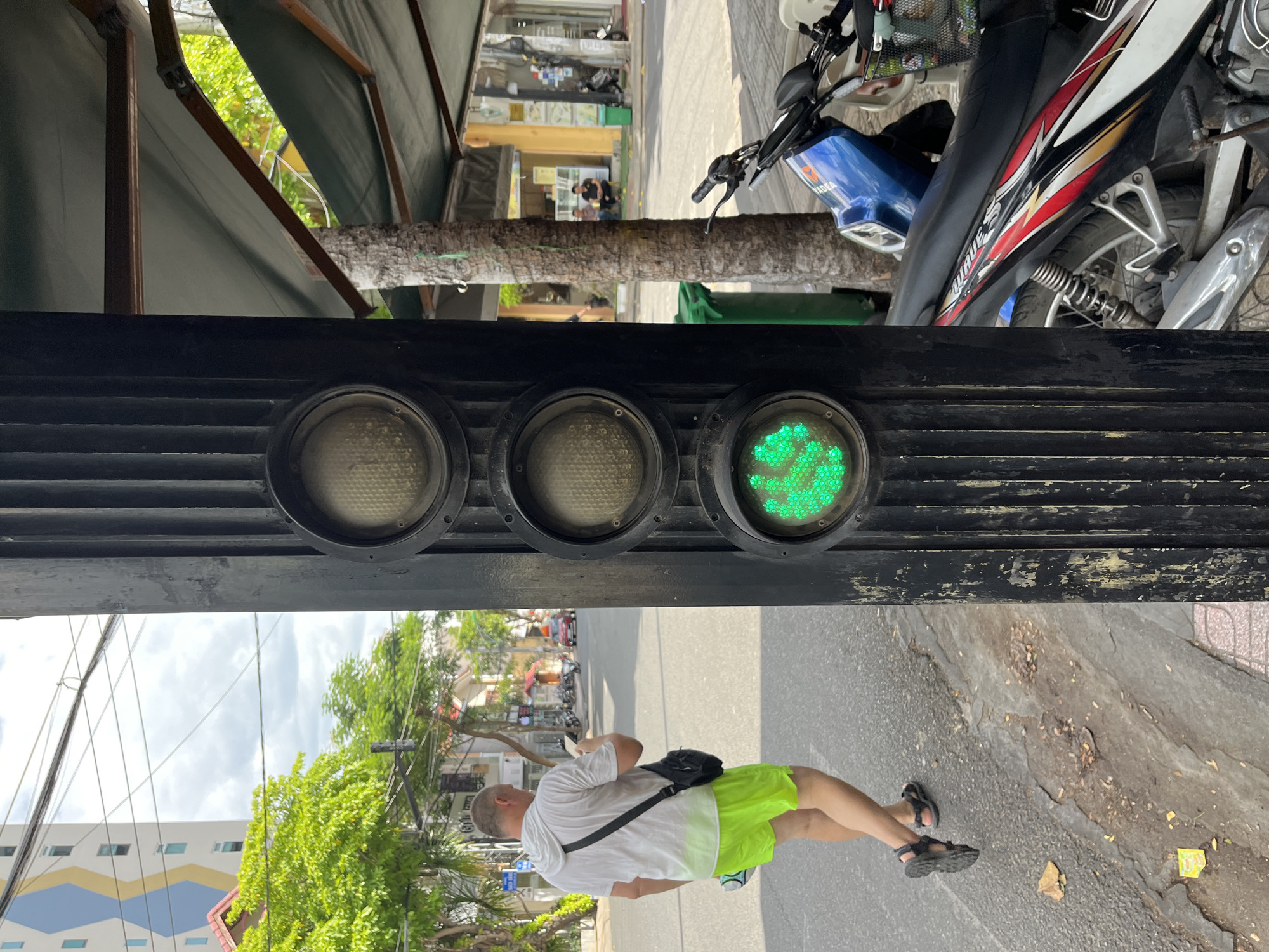 A man wearing a white hoodie and bright green shorts walks away from the camera on a sidewalk next to a traffic light pole in Nha Trang, Vietnam. The traffic light has three circular lenses, with the bottom lens illuminated green. The man is carrying a black crossbody bag and a white object, possibly a phone. In the background, there is a street with parked motorcycles and a red car. Buildings and trees line the street. The sidewalk has visible cracks and debris. The overall scene suggests a typical street view in a Vietnamese city.
