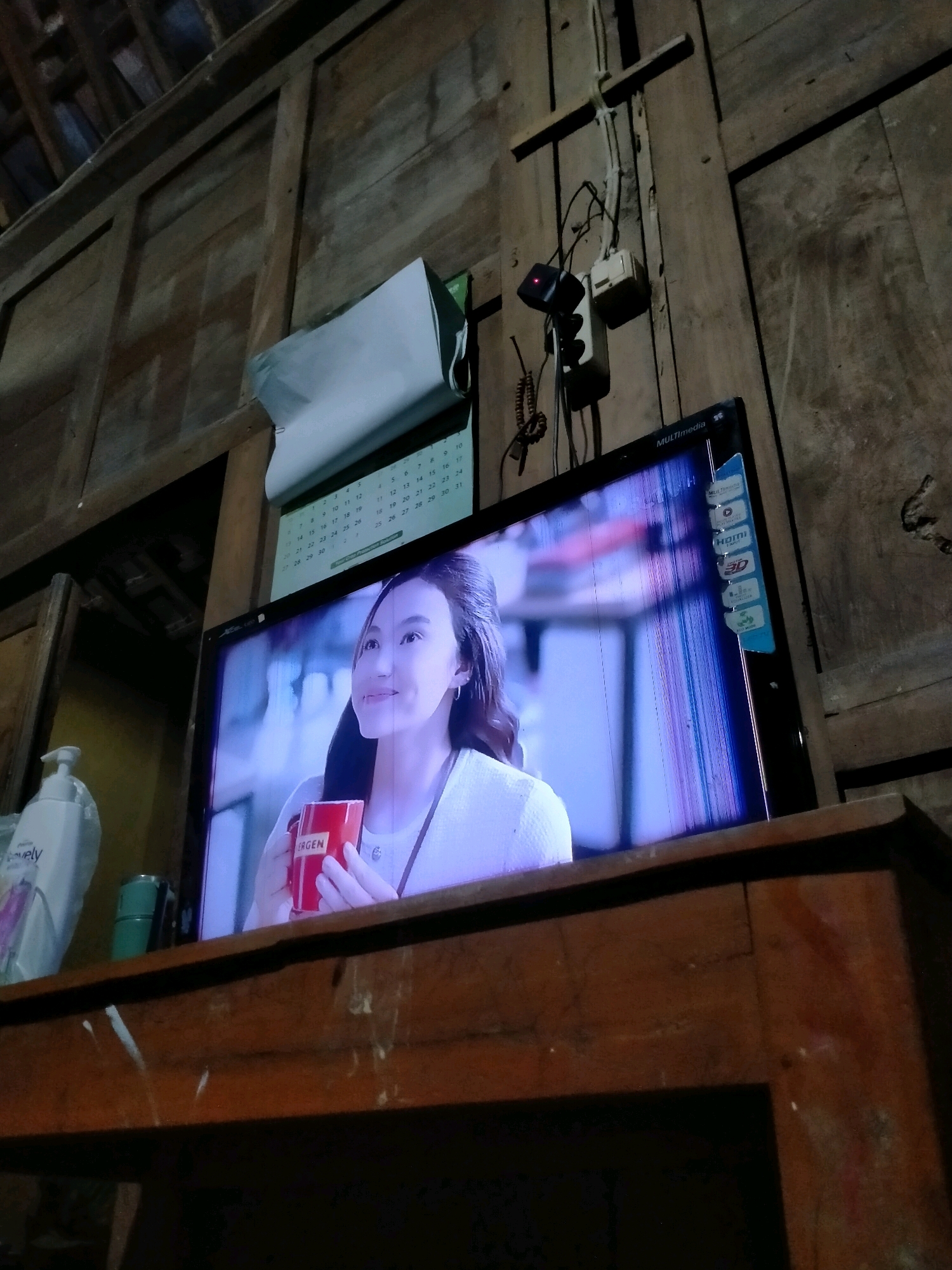 In a room with wooden walls and ceiling, a television screen displays an advertisement for "ERGEN" coffee, featuring a young woman holding a red mug. The television itself appears to be damaged, with visible vertical lines across the screen. On the wall above the television, a calendar is partially visible, along with electrical outlets and wiring. To the left of the television, a bottle of lotion with the brand "Lovely" is present. The scene suggests a domestic setting within Playen, Indonesia, likely a home given the rustic wooden construction.