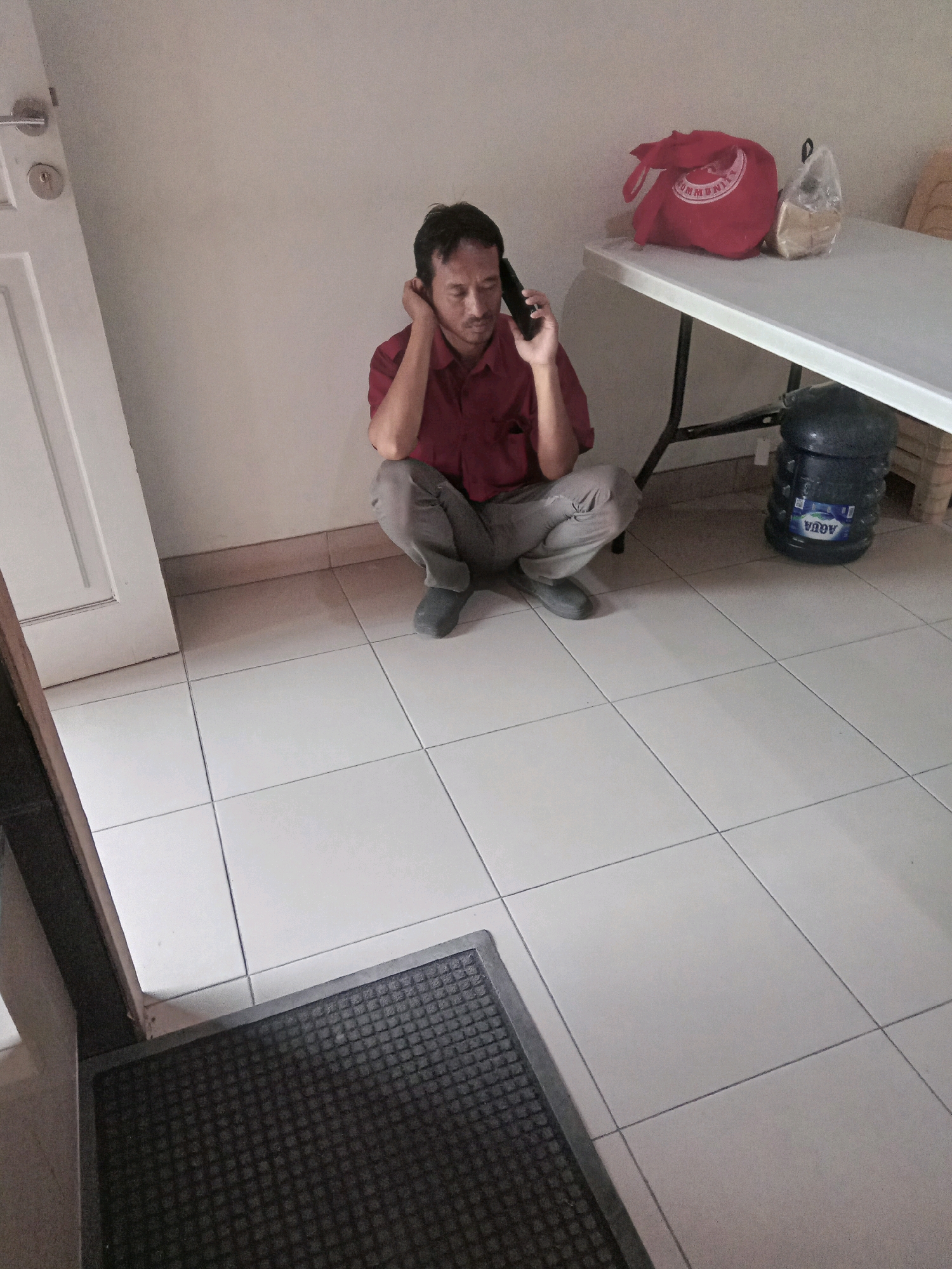 In Surabaya, Indonesia, a man is squatting on a tiled floor, holding a black mobile phone to his right ear. He wears a red collared shirt and grey pants. To his right, a white folding table is set up. On the table, there is a red bag and a clear plastic bag containing light-colored items. Behind the table, a large blue water jug with the label "AQUA" is visible. To the left of the man, a white door with a silver doorknob and lock is slightly ajar. In the foreground, a dark grey doormat with a textured surface is placed on the tiled floor. The walls appear to be a neutral beige color.