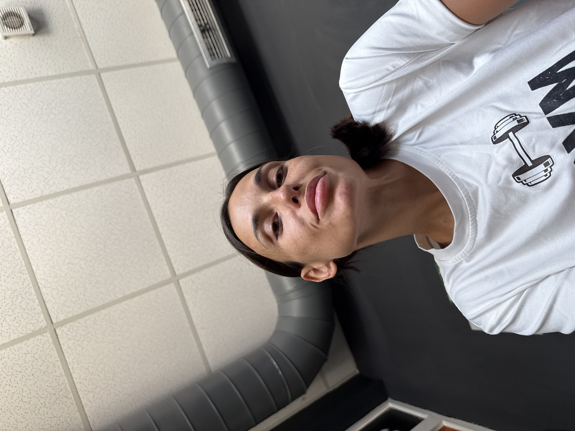 The image is a low-angle, close-up selfie taken indoors. A woman with dark hair pulled back is looking directly at the camera. She is wearing a white t-shirt with a dumbbell graphic and lettering that appears to say "GYM". The background consists of a dark wall, a large grey duct, and a textured white ceiling with fluorescent lighting and a vent. The environment suggests a gym or fitness facility. The location is not specified within the image.