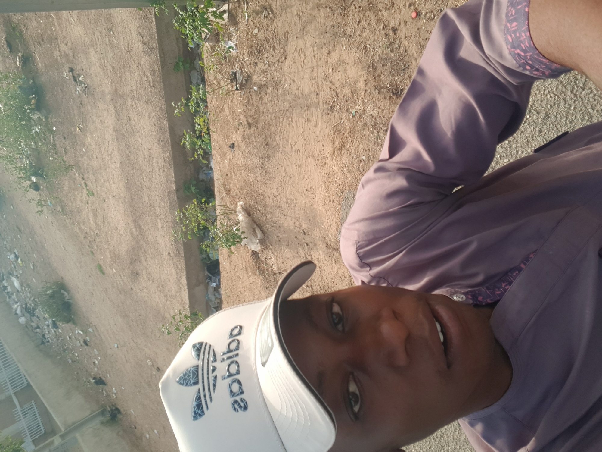 A person wearing an Adidas baseball cap and a light purple collared shirt is shown in a selfie. The background is a dirt area with some scattered vegetation. A concrete curb runs along one edge of the dirt area, and a paved surface can be seen near the person. The location is Dabawa, Nigeria.