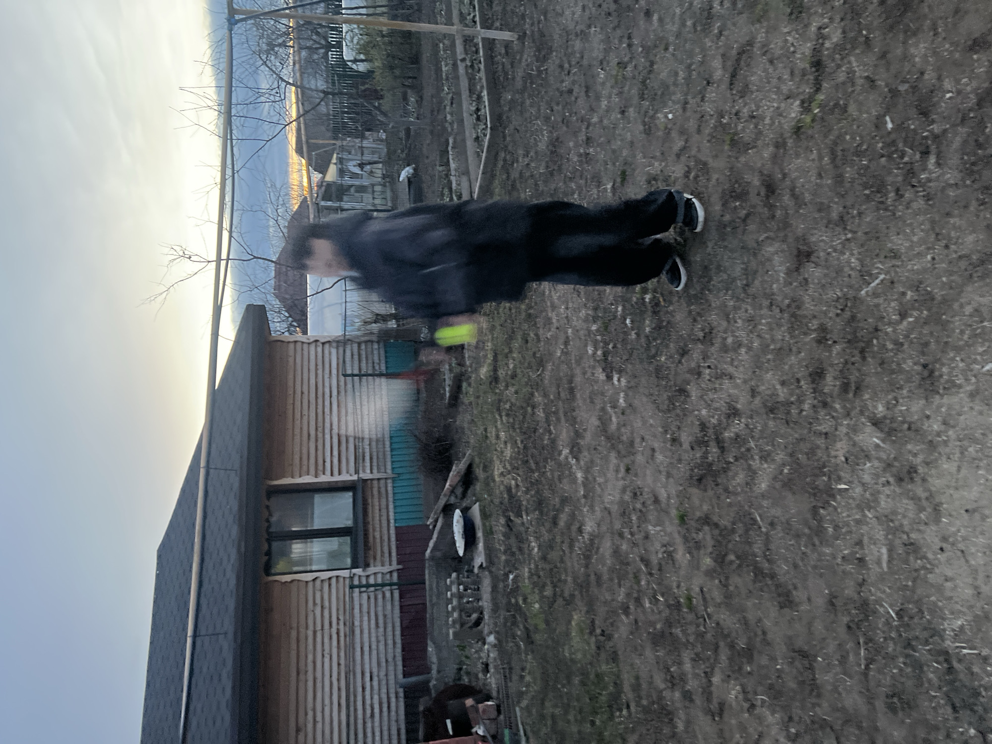 A blurred figure of a person in dark clothing stands on soil or dirt ground. The background consists of a wooden building with a window, bare trees, and the sky. The location is possibly in Votkinsk, Russia.