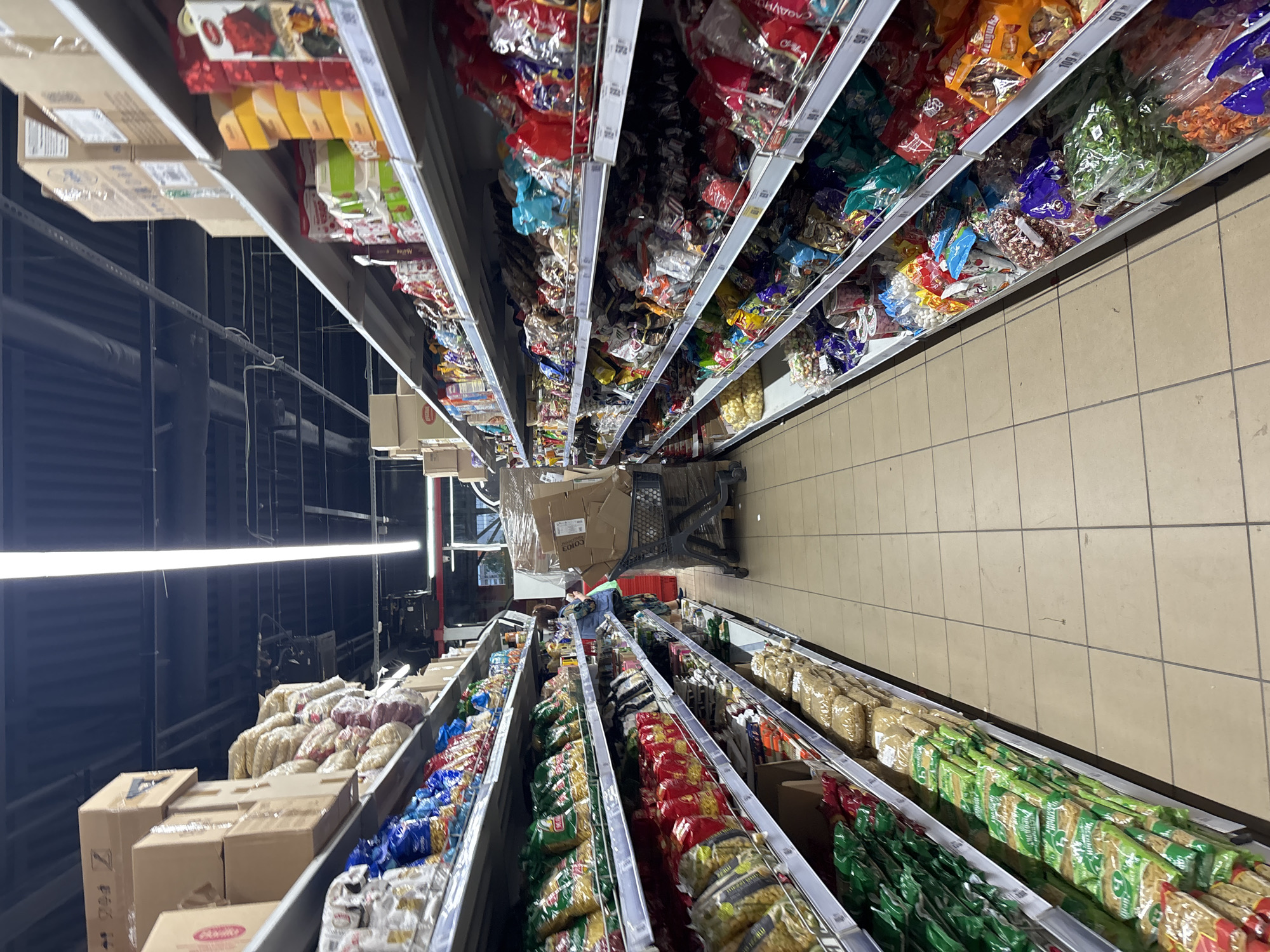 The image shows an aisle in a supermarket, with shelves stocked with food items. The aisle is lined with shelves on both sides, filled with various packaged goods such as pasta, snacks, and other dry food items. The shelves are made of metal and are illuminated by overhead fluorescent lights. The floor is tiled. A shopping cart is visible in the middle of the aisle, with a person pushing it from behind, partially obscured. The person is wearing a patterned dress and carrying a bag over their shoulder. The overall impression is that of a typical grocery store environment, with products neatly arranged on display. The specific location within the city of Unknown, Russia, is not visually identifiable from the image itself.