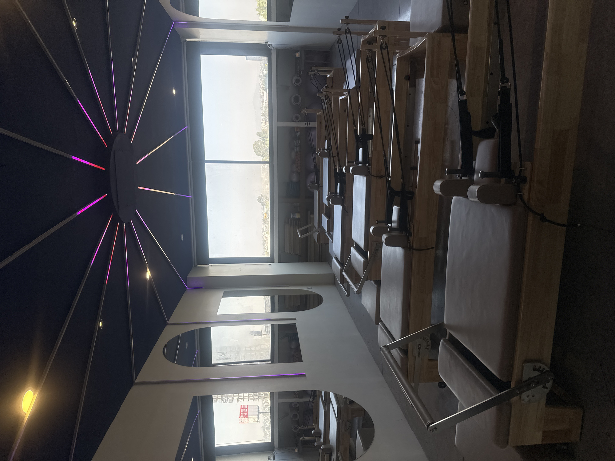 The image depicts a fitness studio in San Bernardino Tlaxcalancingo, Mexico, filled with Pilates reformer machines. Natural light streams in through large windows, offering a view of the surrounding landscape. The studio features a unique ceiling design with a central dark dome and radiating light accents in pink and purple hues. The walls are light-colored, with alternating window and arched mirror panels. The Pilates reformers are made of light wood with beige padding and have black straps and metal components. The studio appears empty, with no visible people engaging in activities.