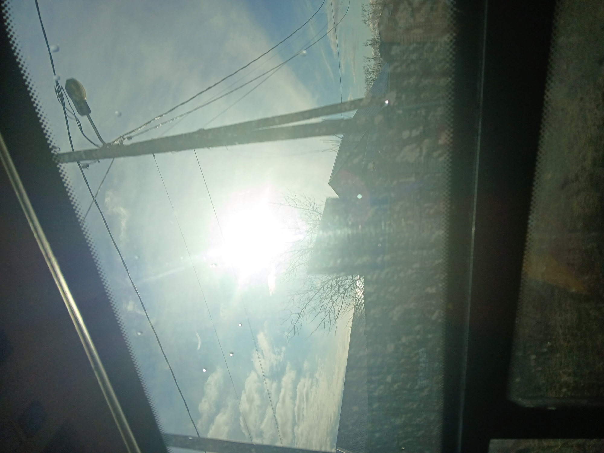 The image shows a view looking up through a sunroof, or a similarly angled window of a vehicle. The sky is visible, with bright sunlight obscured by thin clouds. Utility poles and wires are visible, as is a bare tree. The location is likely Berezniki, Russia, based on the prompt. The interior of the vehicle frames the view, with portions of the vehicle's roof and interior visible along the edges. The glass of the sunroof has some water spots and other minor imperfections.