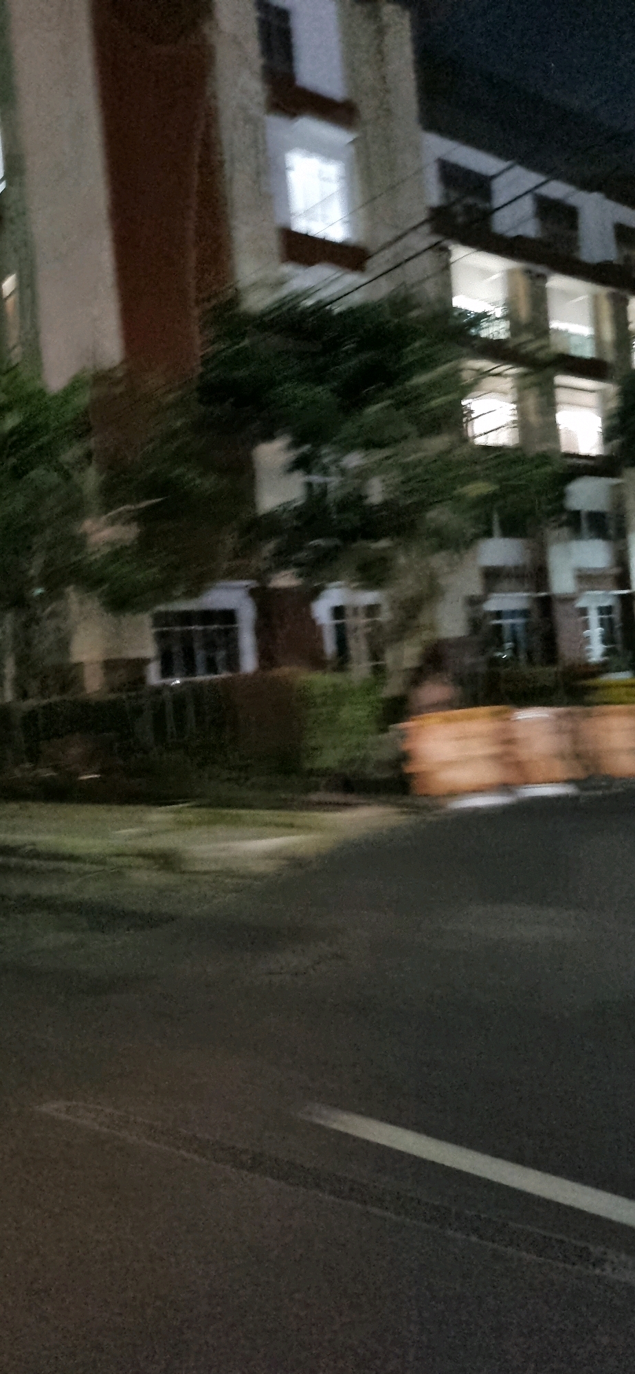 The image depicts a blurred view of a building at night, likely in Denpasar, Indonesia. The building appears to be multi-story with visible windows, some of which are illuminated. There is a dark road in the foreground with tire marks and a white line. Greenery, possibly trees or hedges, is visible in front of the building. In the middle ground, there are orange barriers that appear to be traffic barriers. The overall impression is of a street scene at night, possibly taken from a moving vehicle, resulting in motion blur.