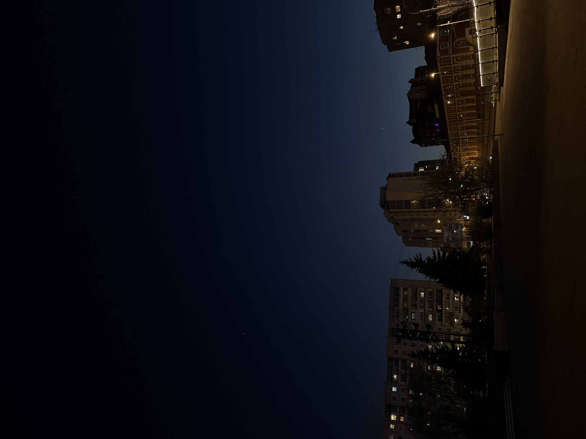 The image shows a night view of Krasnoyarsk, Russia. On the right side, buildings are visible, including a brick building with ornate decorations and a more modern apartment building. Streetlights illuminate the buildings and the sidewalk. The left side of the image is a dark blue sky.
