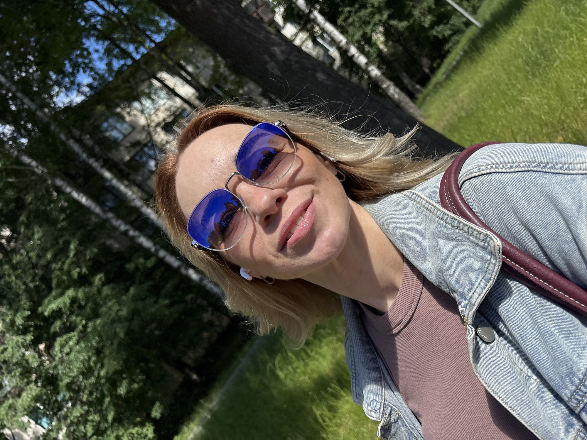 A woman with blonde hair and sunglasses takes a selfie outdoors, with a blurred background of trees and buildings. She is wearing a denim jacket over a rust-colored shirt and appears to have white wireless earbuds in her ears. A burgundy strap from a bag is visible over her shoulder. The scene suggests a sunny day in a park or residential area, possibly with a city skyline in the distance. There is no specific indication of the location being Saint Petersburg, Russia, beyond the provided prompt.