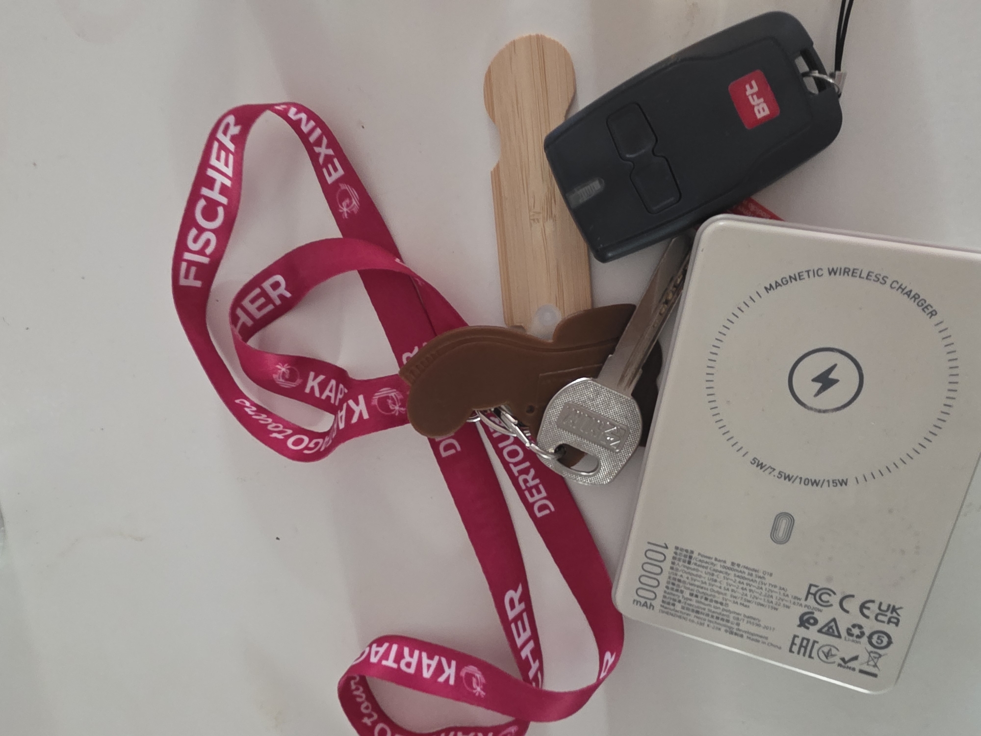 The image shows a collection of personal items laid out on a white surface. In the center, a cluster of keys with various attachments is visible. One key has a silver tag with text that is partially obscured but appears to be a brand name. Attached to the keys is a brown, somewhat abstract-shaped charm. A bright pink lanyard with white text is draped around the keys and charm. The text on the lanyard includes the words "FISCHER," "KARTAGO TOURS," and "EXIM." Also in the image are a white rectangular power bank with markings indicating "100000 mAh" and "MAGNETIC WIRELESS CHARGER," and a black remote control with a red "BfT" logo. A small, light-colored wooden object with a curved end is positioned beneath the keys. The white surface appears to be a tabletop or counter. There are no people or animals in the image. The scene appears to be an indoor setting, with the white surface suggesting a clean, neutral background. The lighting is even, indicating a well-lit environment.