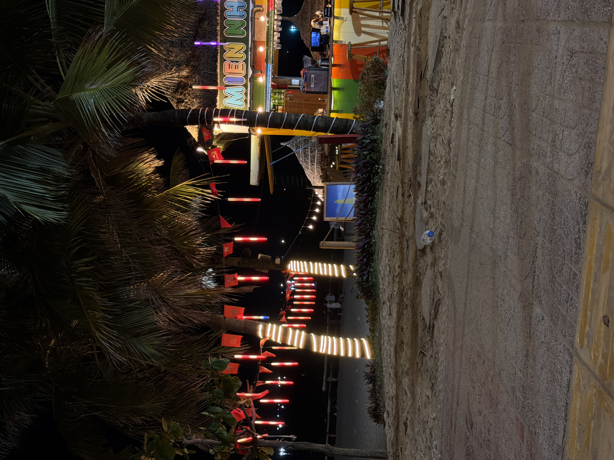 The image depicts a nighttime scene on a beach in Da Nang, Vietnam, with a bar and palm trees decorated with lights and flags. In the foreground, there is a sandy beach with some paving stones. Further back, a bar is set up with a colorful facade and stools. Two palm trees are prominently featured, wrapped in string lights. Red flags, some with yellow stars, are hung between the trees, suggesting a Vietnamese context. The overall ambiance is festive and tropical.
