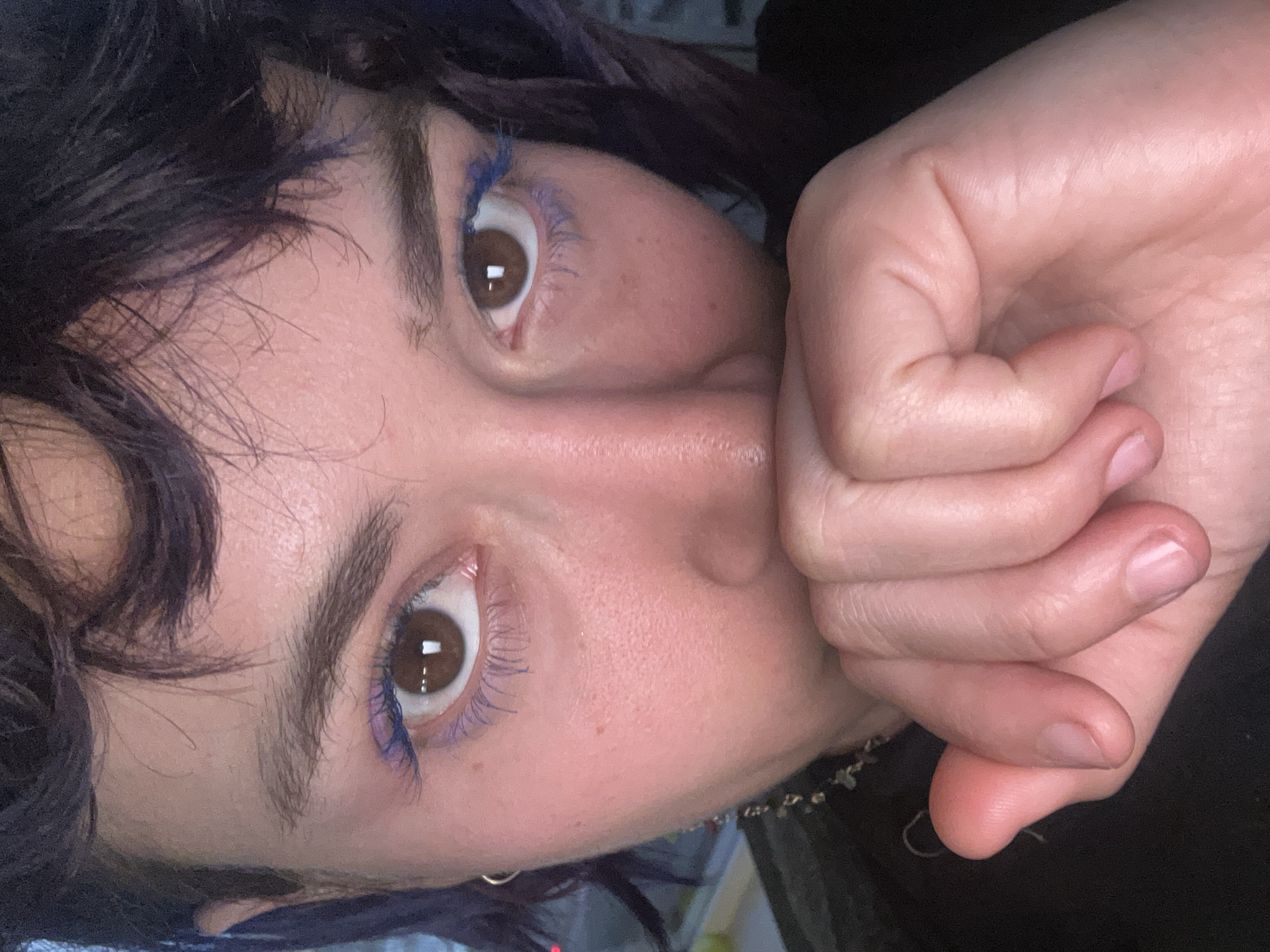 This image depicts a close-up, eye-level portrait of an individual in an indoor setting within the city of Los Angeles, United States of America.
The subject is a person, likely an adult female, with a direct gaze fixed upon the camera. Their facial features include brown eyes with visible circular reflections in the pupils, indicating an artificial light source or window. Both upper and lower eyelashes are coated with bright blue mascara. Dark, natural eyebrows are visible, along with short, wavy, dark hair exhibiting a purplish tint, which frames the face. The skin complexion is fair, displaying visible pores and minor blemishes. A small silver hoop earring is present on the subject's left earlobe.
The individual's right hand is raised to the lower portion of their face, with fingers curled and pressed against the mouth and chin area, effectively obscuring these features. The fingernails are short and unpolished. Visible on the left side, beneath the ear, is a metallic chain, possibly a necklace, featuring dark, irregular elements. The subject appears to be wearing dark-colored clothing, visible as a collar or neckline on the left.
The background is heavily blurred and out of focus, revealing diffuse light sources. This suggests an indoor environment, possibly at night or in a dimly lit room, with indistinct lights from either outside a window or within the room itself. A faint red light source is discernible in the blurred background on the left.