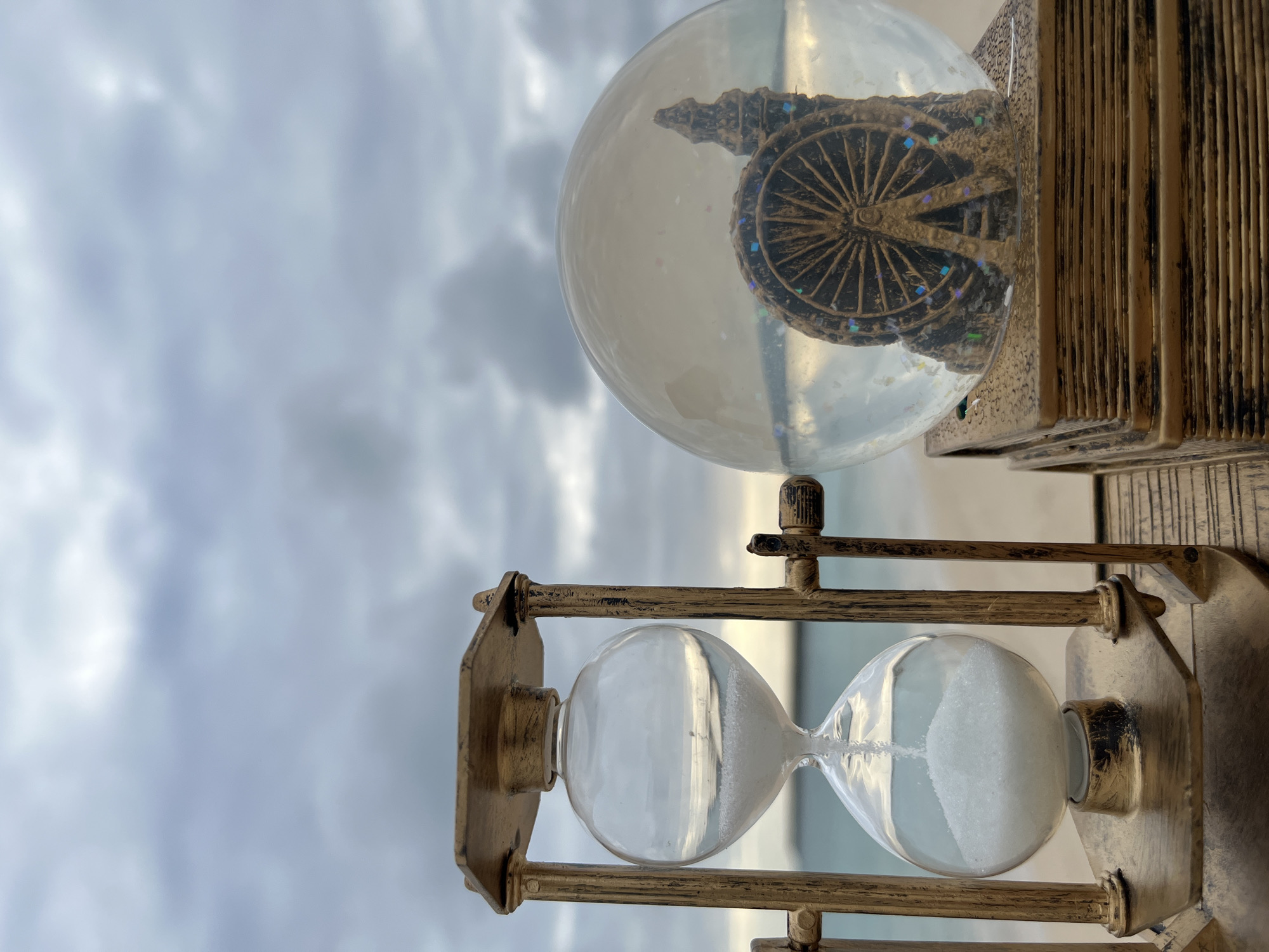 The image shows a close-up of a golden hourglass next to a snow globe on a weathered wooden surface. The hourglass has white sand flowing from the top bulb to the bottom bulb. The snow globe contains a miniature Ferris wheel and what appears to be a building or monument, set against a backdrop of a cloudy sky and a beach. The sea is visible beyond the beach. The location is not specified as Kuta, Indonesia.