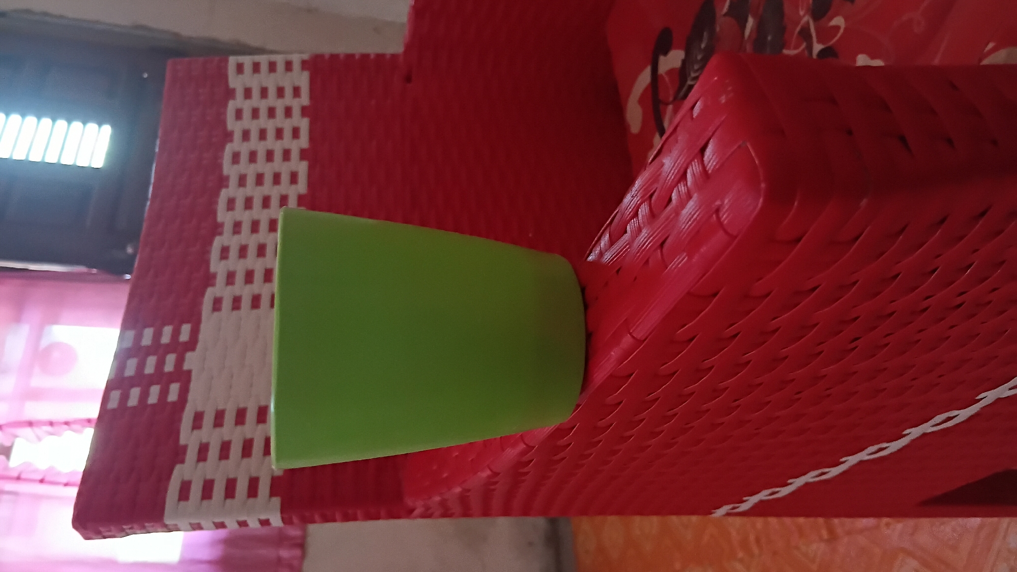 A green plastic cup rests on the armrest of a red woven plastic chair. The chair has a white woven pattern on its back. In the background, sheer pink curtains are visible, and a window with blinds is to the right. The scene appears to be indoors, possibly in a residential setting in Prabumulih, Indonesia, given the context provided.