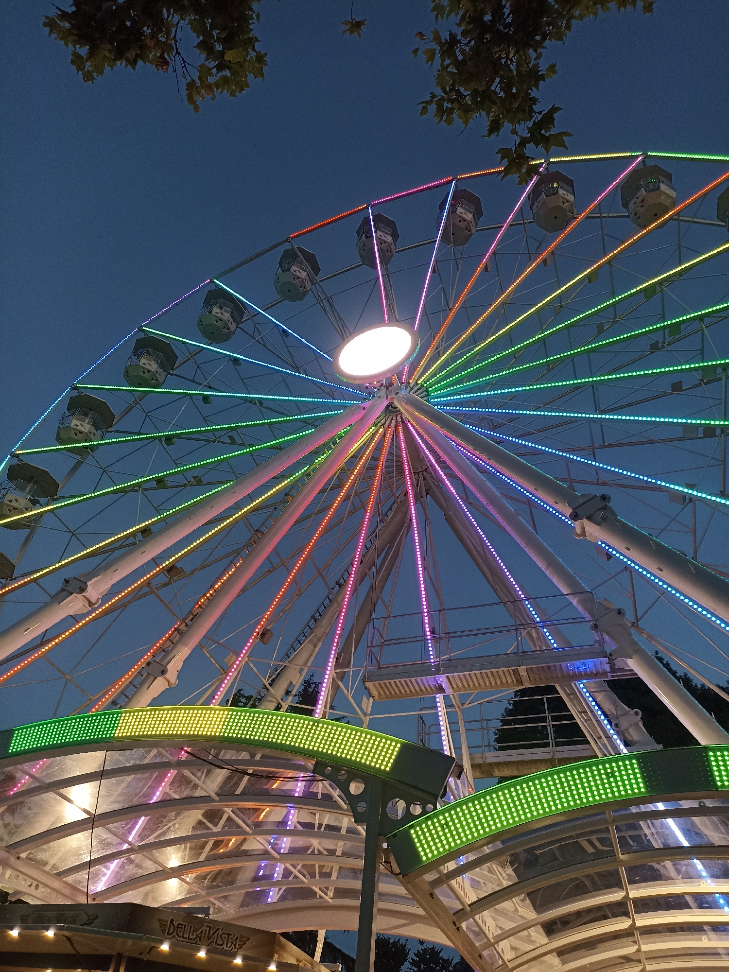 The image shows a Ferris wheel at dusk, illuminated by colorful lights. The Ferris wheel is a prominent feature, with its white structure and multicolored LED lights that cycle through green, yellow, orange, pink, and blue. The cabins are visible, suspended from the wheel. In the foreground, there is a covered structure with green and yellow LED lighting. Above the Ferris wheel, a branch with autumn leaves is visible against the dark blue sky. The scene is likely from an amusement park or a fairground. Based on the provided information, this Ferris wheel is located in Burgas, Bulgaria. The overall atmosphere is festive and illuminated.