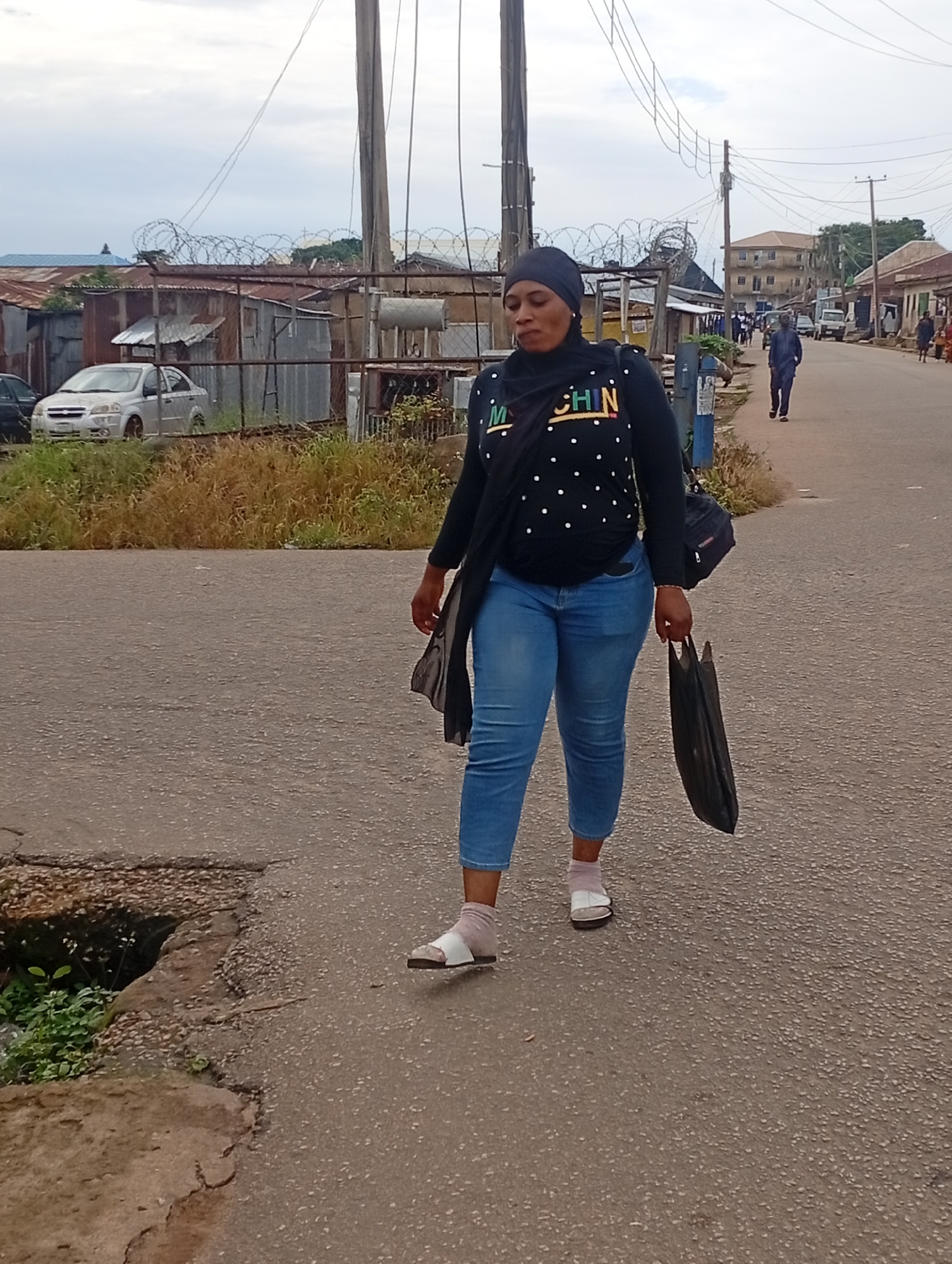 A full-body, eye-level, medium shot captures a woman walking on an asphalt road in an outdoor urban setting. She is positioned in the left foreground, facing right and looking forward. The woman wears a black hijab, a black long-sleeved shirt with white polka dots and "MCHIN" text in colorful letters, and light blue jeans. She carries a black purse on her right shoulder and a black plastic bag in her right hand. Her feet are adorned with white socks and white sandals. To the left of the woman, a pothole is visible in the road, with green plants growing from within. In the background, a silver car is parked behind a fence with barbed wire. Several buildings, some appearing to be made of corrugated metal and others of brick, line the street, along with utility poles and wires. Other pedestrians and vehicles are present further down the road, suggesting a busy street. The sky is overcast and cloudy.