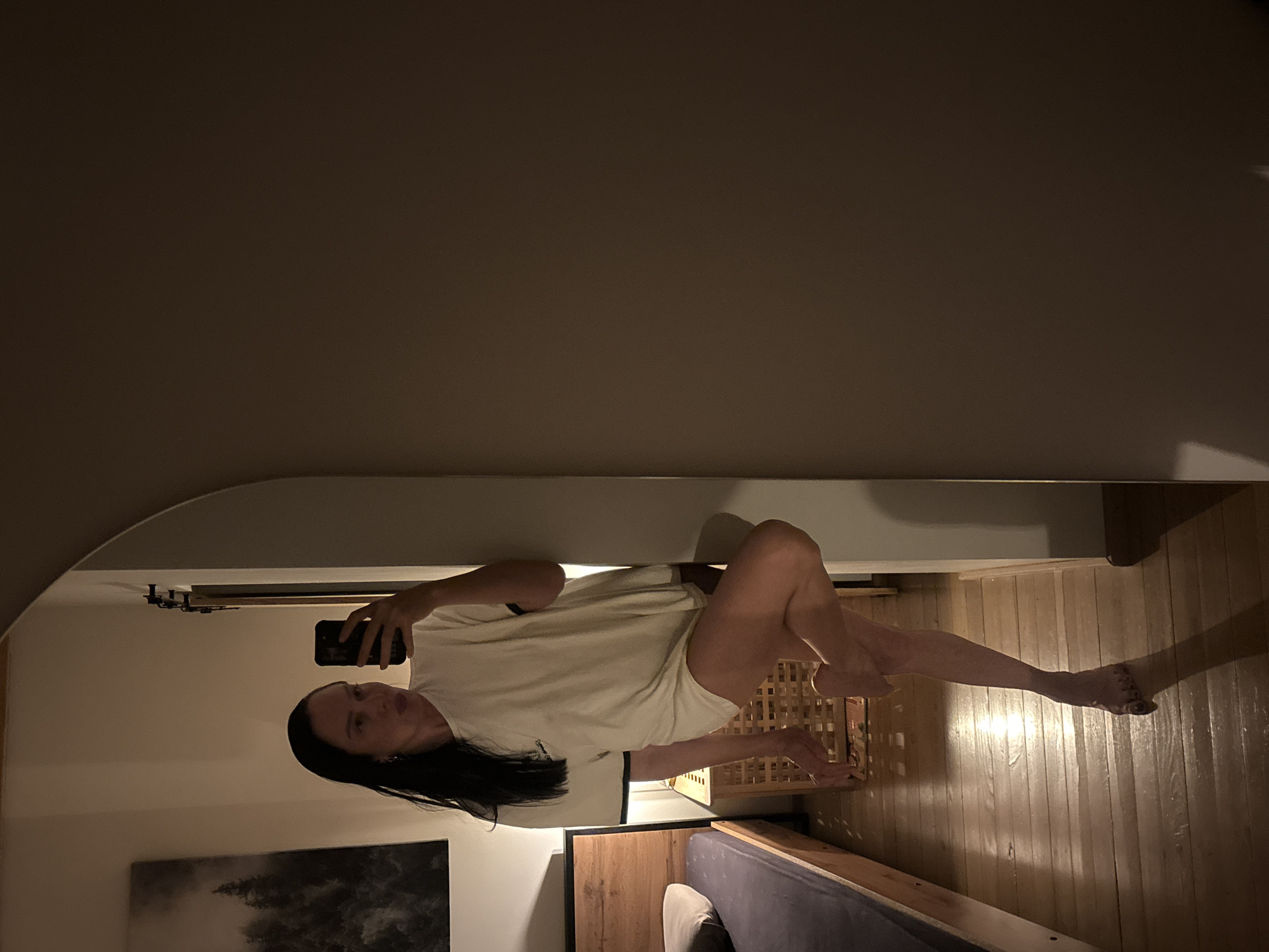A woman is seen in a reflection from a mirror. She is holding a phone and appears to be taking a selfie. She is wearing a white t-shirt. Her location is a bedroom. A painting can be seen on the wall. A bed and a wooden container are also visible. The location is not in Chaikovsky, Russia.