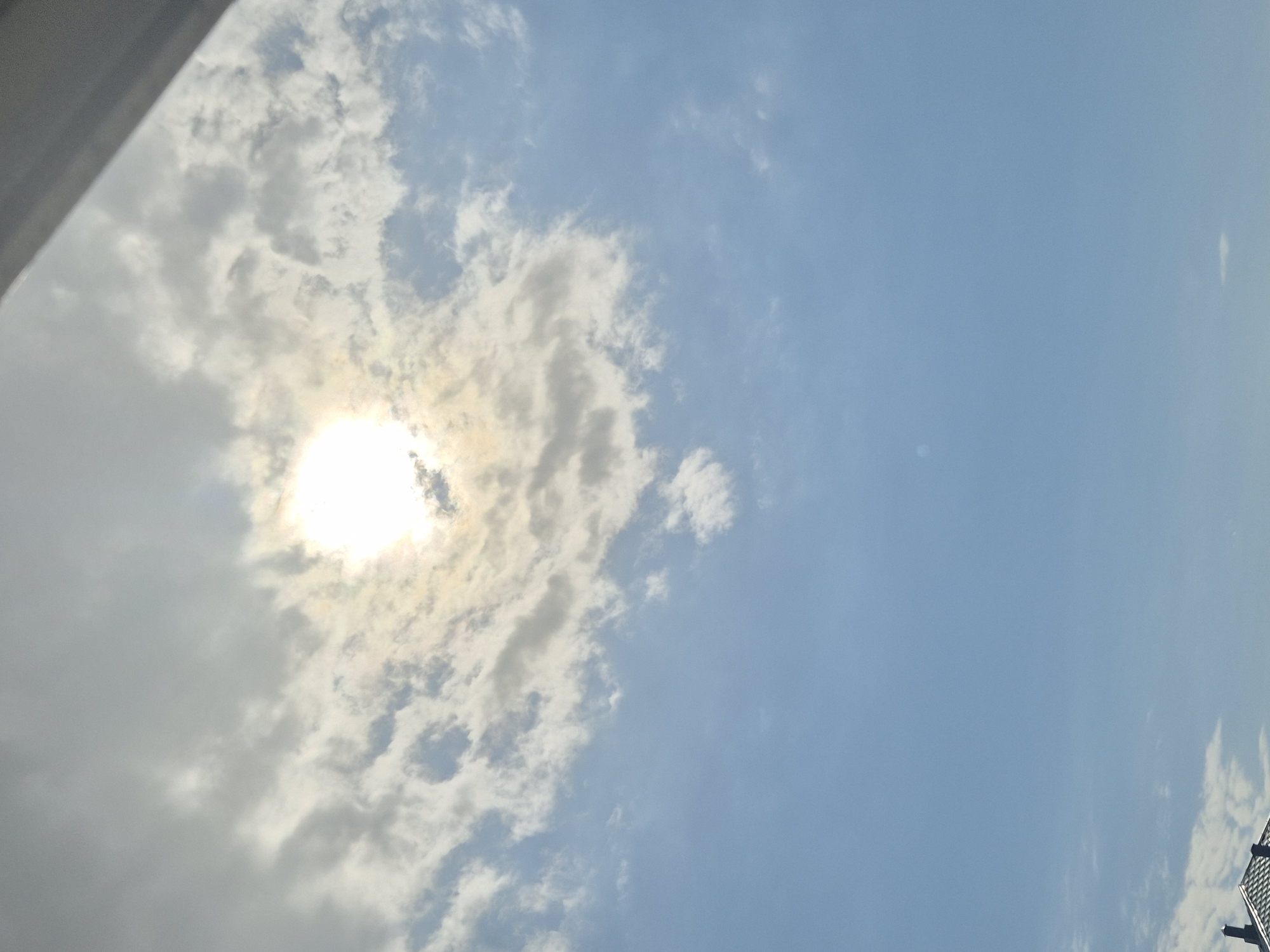 The image shows a sky with a mix of clouds and clear blue areas. The sun is visible, obscured by a layer of clouds, creating a bright spot. Based on the limited visible information, and the request that it be located in Kokshetau, Kazakhstan, it is reasonable to suggest the picture was taken outdoors in that city.