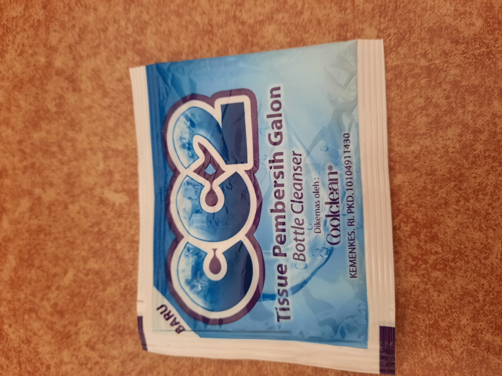 A single packet of "CC2 Bottle Cleanser" tissue is pictured lying on a textured, brown surface, likely a table. The packet has a blue and white design with the brand name "CC2" prominently displayed in large, stylized letters. Below the brand name, the text "Tisu Pembersih Galon Bottle Cleanser" is visible. Further down, it states "Dikemas oleh: Coolclean" and includes registration information "KEMENKES. RI. PKD. 10104911430". The packet appears to be new, indicated by the word "BARU" in the top left corner. The image is a close-up shot of the packet, with the background slightly out of focus. There are no people, animals, or any other identifiable objects in the frame besides the packet and the surface it rests on.