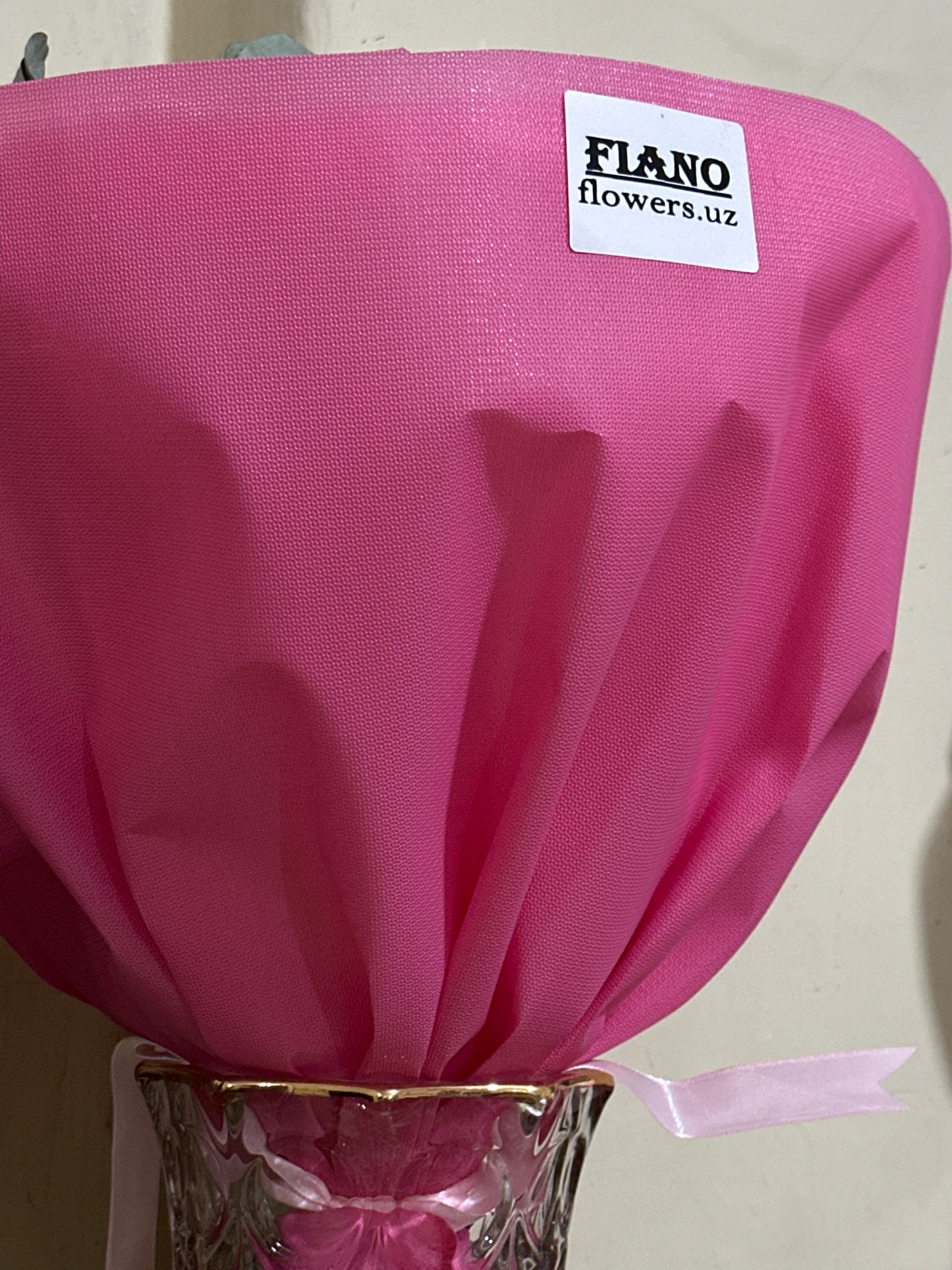 A close-up shot focuses on a bright pink bouquet of flowers wrapped in decorative paper. A small white sticker with black lettering is affixed to the paper, reading "FLANO flowers.uz". A delicate light pink ribbon is tied around the base of the bouquet where it meets a clear glass vase with a gold rim. The vase appears to be filled with more pink flowers. The background is a neutral, textured wall. There is no indication of the city of Salar, Uzbekistan within the image itself.