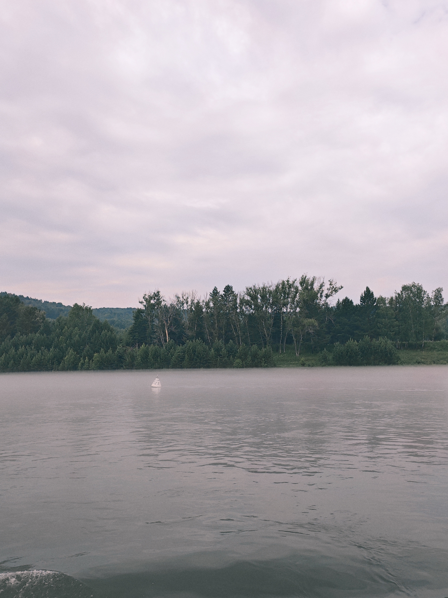 A body of water, likely a river, is shown with a dense forest lining the distant bank. The sky is overcast and appears to be a light grey with some hints of pink. A white buoy, labeled with the number "23", is floating on the water. A light mist or fog hovers just above the water's surface, particularly near the shore. The water itself is calm with subtle ripples. The scene is tranquil and appears to be in the early morning or late evening. The location is Atamanovo, Russia, a small village situated on the Yenisei River.