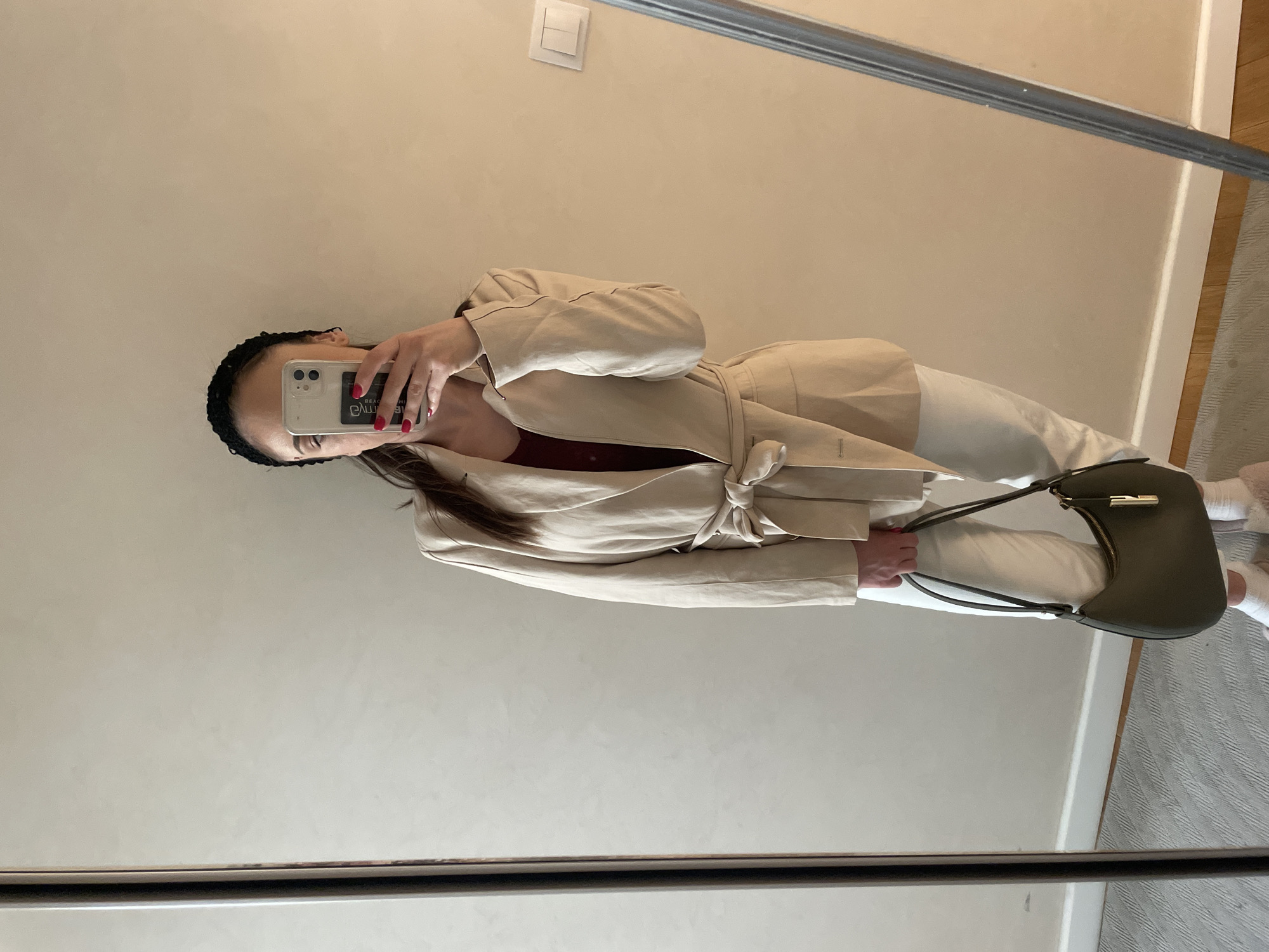 A person stands indoors, taking a mirror selfie. They wear a beige belted blazer, white pants, and carry a green handbag. The background is a plain, light-colored wall. The setting is not identifiable as Varna, Bulgaria from the image alone.