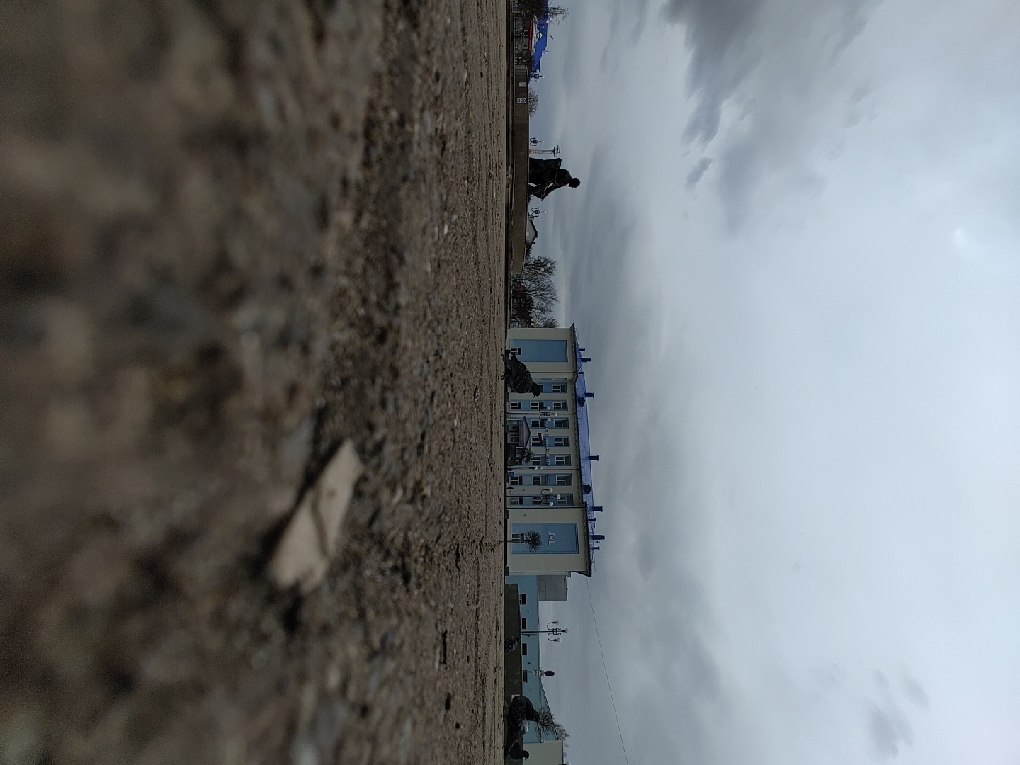 An outdoor, low-angle shot captures an overcast day in what appears to be an urban or semi-urban setting in Chaikovsky, Russia. The foreground, occupying a significant portion of the lower frame, is blurry and consists of rough, gravelly ground. A small, light-colored piece of debris is visible within this blurred area.
In the midground, towards the left, a pigeon walks on the ground. To the right of the pigeon is a light-colored, multi-story building featuring distinctive blue accents on its roofline and window frames. A prominent, vertically oriented sign on the side of this building clearly displays the Cyrillic text "ТОТ". Further to the right, positioned on a slightly elevated platform or wall, a dark, silhouetted figure of a person is visible, appearing to be sitting or crouching.
The background is dominated by a vast, overcast sky, filled with shades of gray clouds, indicating cloudy or gloomy weather. Along the distant horizon, faint outlines of bare trees and other structures can be discerned. The overall impression is one of a quiet, everyday scene under a muted sky.