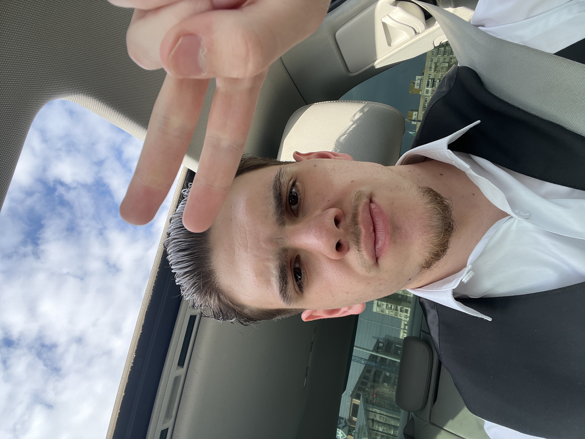 A young adult male is depicted in a selfie taken from the interior of a vehicle in Sofia, Bulgaria. The subject, positioned centrally, has short, slicked-back dark hair exhibiting some lighter strands, dark eyes, a light mustache, and a goatee. He is dressed in a white collared shirt and a black vest, secured by a grey seatbelt across his chest. His right hand is raised, forming a "peace" (V) sign with the index and middle fingers. The car's interior includes a grey headliner and an open sunroof exposing a blue sky with scattered white clouds. Through the tinted rear side windows, an urban landscape is visible. To the left, several multi-story modern buildings are under construction, with a crane partially visible. To the right, an older, multi-story residential building with numerous windows is present. The environment is consistent with an urban district in Sofia featuring a mix of new construction and established residential architecture.