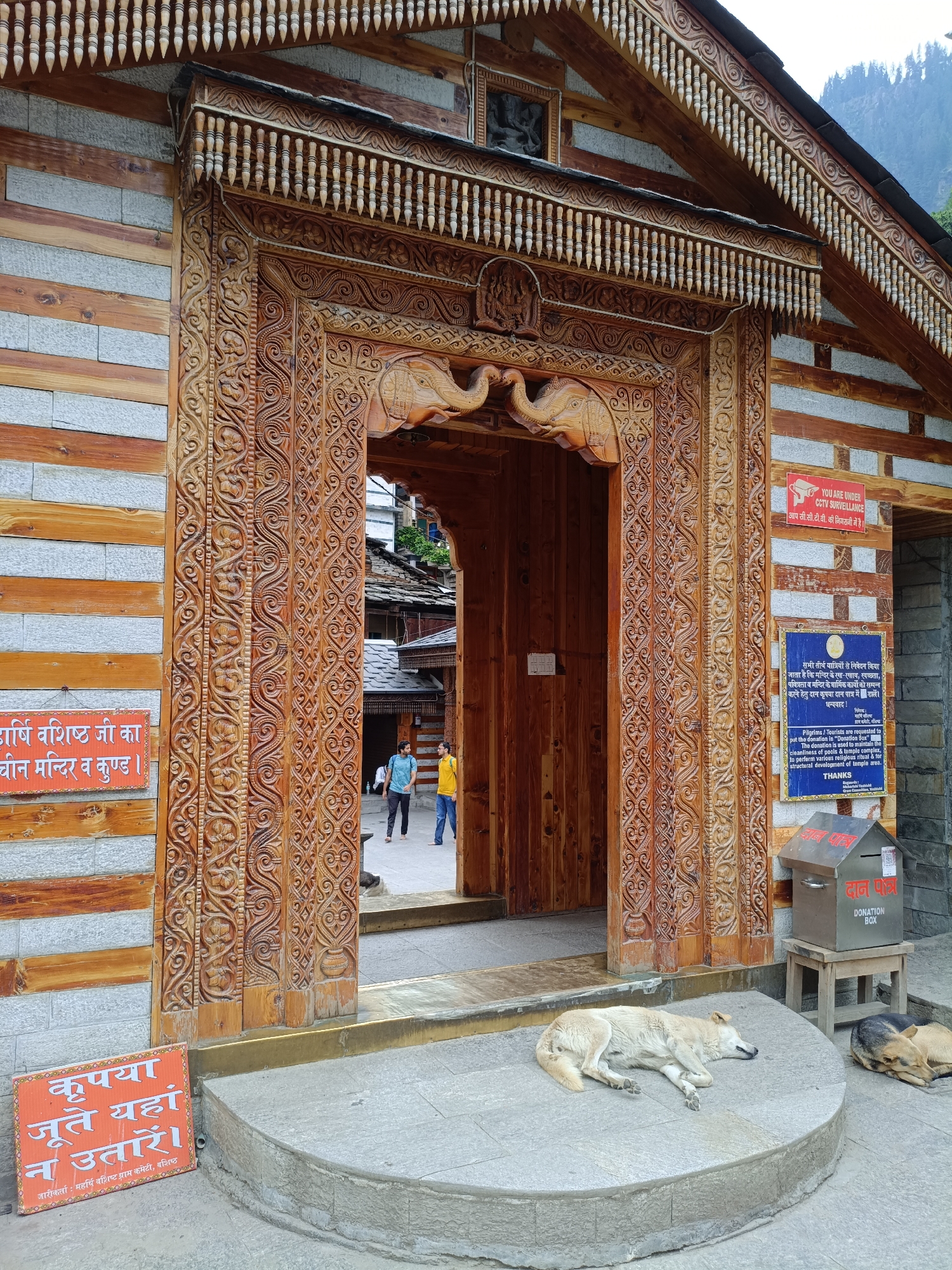 The image shows the entrance to a building in Vashisht, India, featuring ornate wooden carvings. The building's exterior is constructed with stone and wood. In the foreground, two dogs are sleeping on a stone platform. One dog is light-colored and lying on its side, while the other is darker and curled up. To the left of the entrance, an orange sign with Hindi text reads "Please do not leave your shoes here." To the right, a blue sign with Hindi and English text requests pilgrims and tourists to maintain cleanliness and make donations. Above the blue sign, a red and white sign states "You are under CCTV surveillance." Two men are walking through the wooden doorway in the background. The doorway itself is elaborately carved with floral patterns and elephant motifs. A donation box is placed on a wooden stool to the right of the entrance. The overall scene suggests a temple or a historical building in a public area.