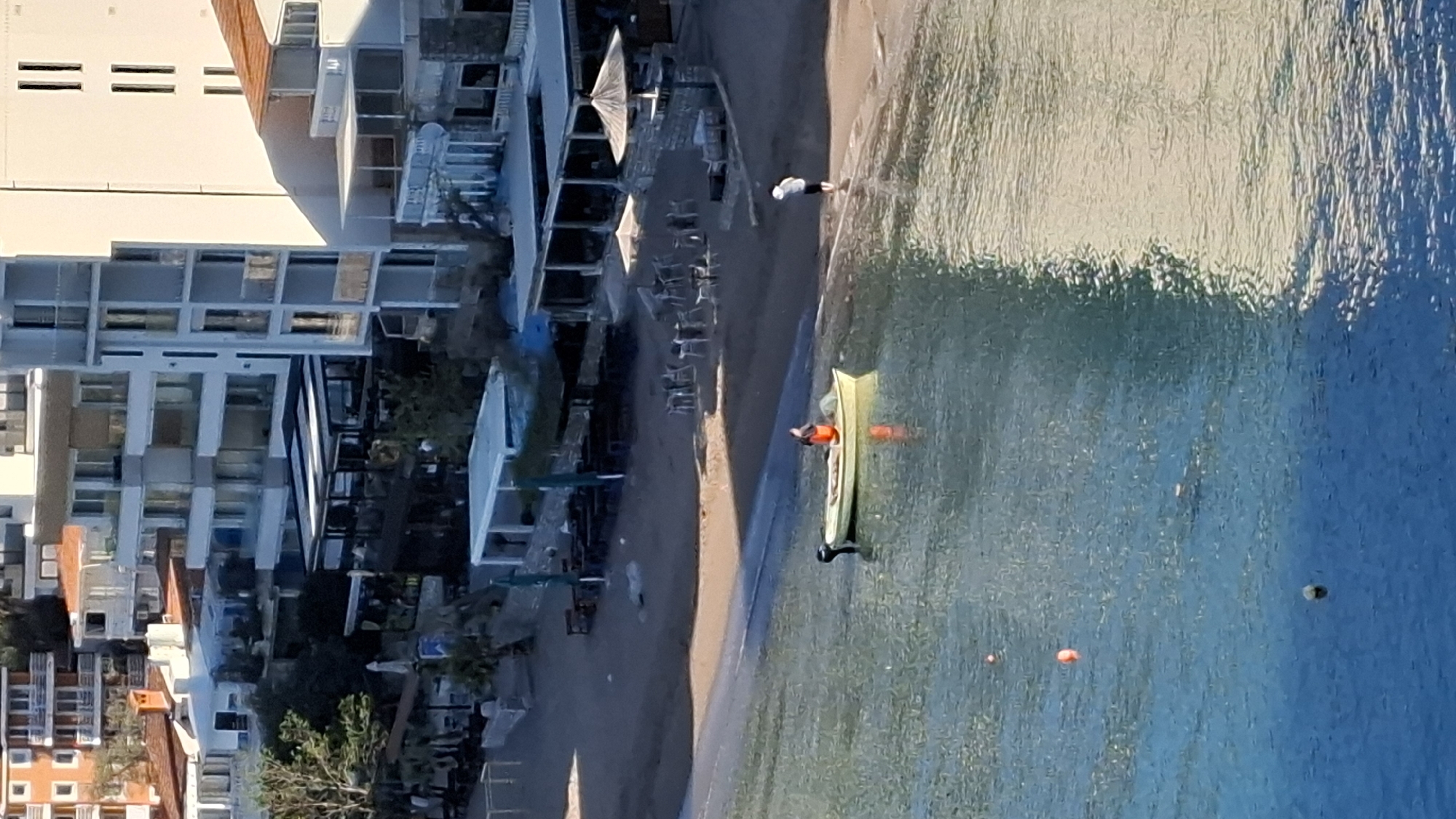 The image depicts a coastal scene with a beach and buildings in Budva, Montenegro. Along the shore, there is a beach with lounge chairs under umbrellas. Buildings, predominantly white and multi-storied, line the coast. The sea is a gradient of light to dark blue. A single kayak with a person is on the water. Another individual is seen walking along the beach.