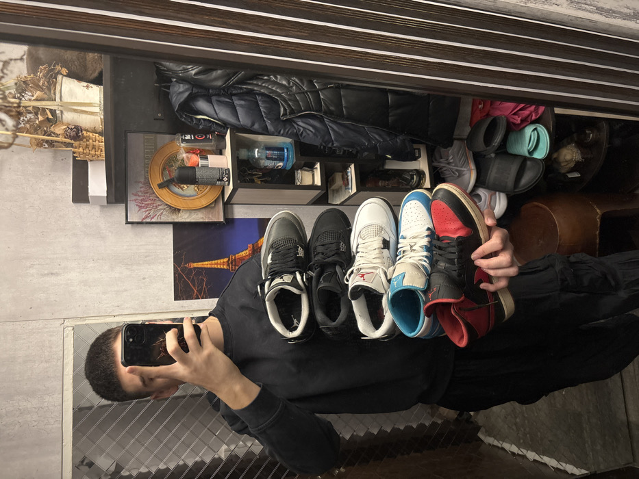 The scene depicts a male individual taking a mirror selfie inside what is identified as a residential interior in Sofia, Bulgaria. The individual, visible from the mid-torso down and partially obscured by a smartphone held to capture the reflection, wears a dark long-sleeved top and dark cargo-style pants. His left hand is extended forward, vertically stacking five distinct pairs of sneakers, while his right hand holds the smartphone. The sneakers, from top to bottom, are: a dark gray/black pair (likely Air Jordan 4), a black pair (likely Air Jordan 4), a white pair with red accents (likely Air Jordan 4), a white and light blue low/mid-top pair (likely Air Jordan 1), and a black and red high-top pair (likely Air Jordan 1). The background, reflected in the mirror, shows a textured light gray wall. To the right, a shelving unit displays various items including decorative baskets with dried flowers, a framed picture depicting lavender, a round framed object, several aerosol cans/bottles (deodorant, hairspray), and a dark puffer vest hanging. Lower shelves contain additional footwear, including sneakers and slides/sandals, next to a dark wooden stool. A poster featuring the Eiffel Tower at night is visible on the wall behind the individual's left shoulder. The mirror itself has a dark brown frame with vertical lighter stripes on its right edge.
