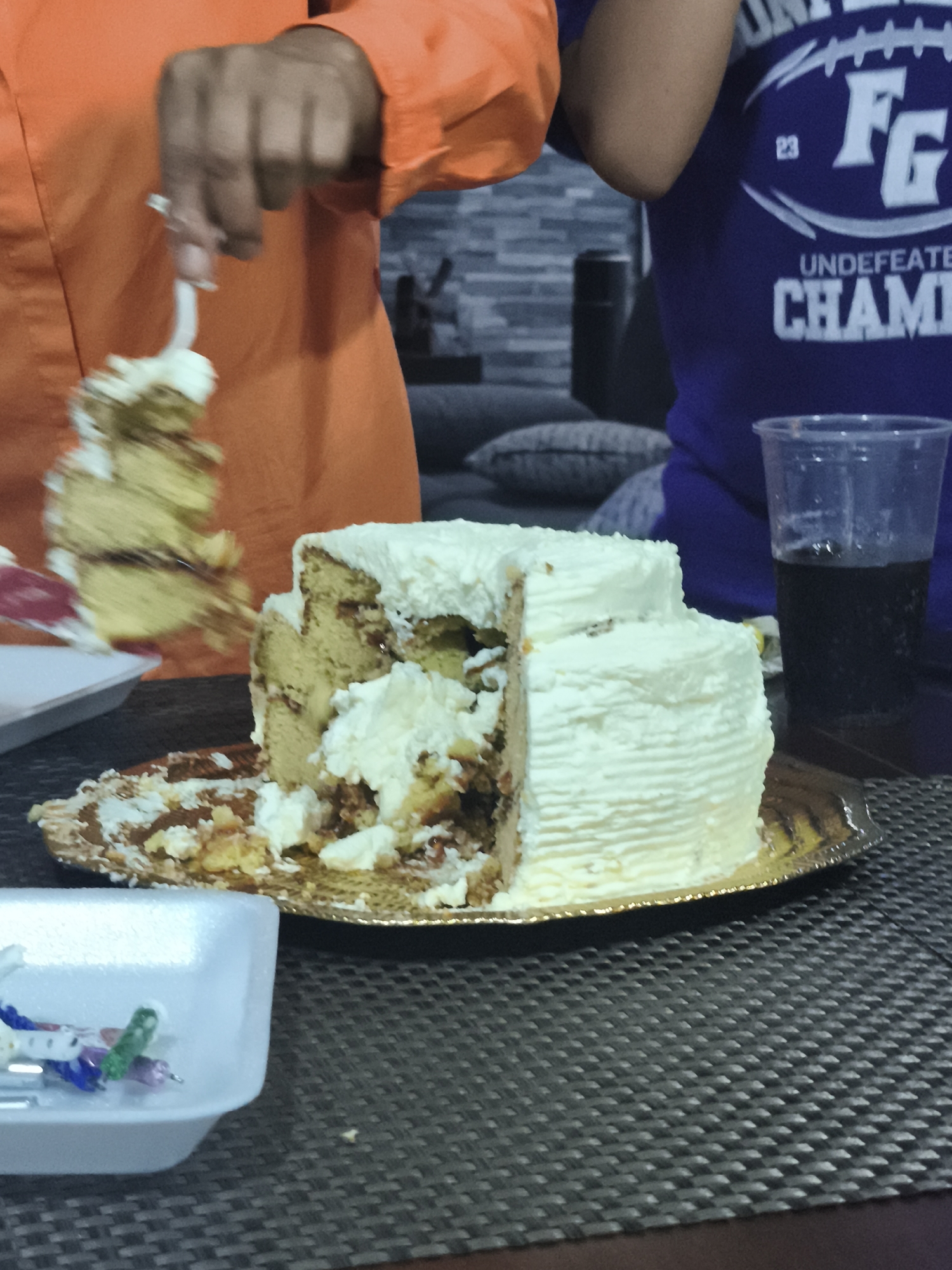 A photograph taken in a residential setting features a two-tiered cake with white frosting on a gold-colored platter. A slice of cake is being lifted from the cake by a person wearing an orange shirt. To the right, a person wearing a purple shirt with the letters "FG" and the words "UNDEFEATED CHAMPS" on it is also visible. In the foreground, a white rectangular container holds small plastic forks or spoons. A plastic cup filled with a dark liquid is also visible near the cake. The background shows a textured gray wall and a gray sofa with pillows. The location is identified as Mexico City, Mexico.