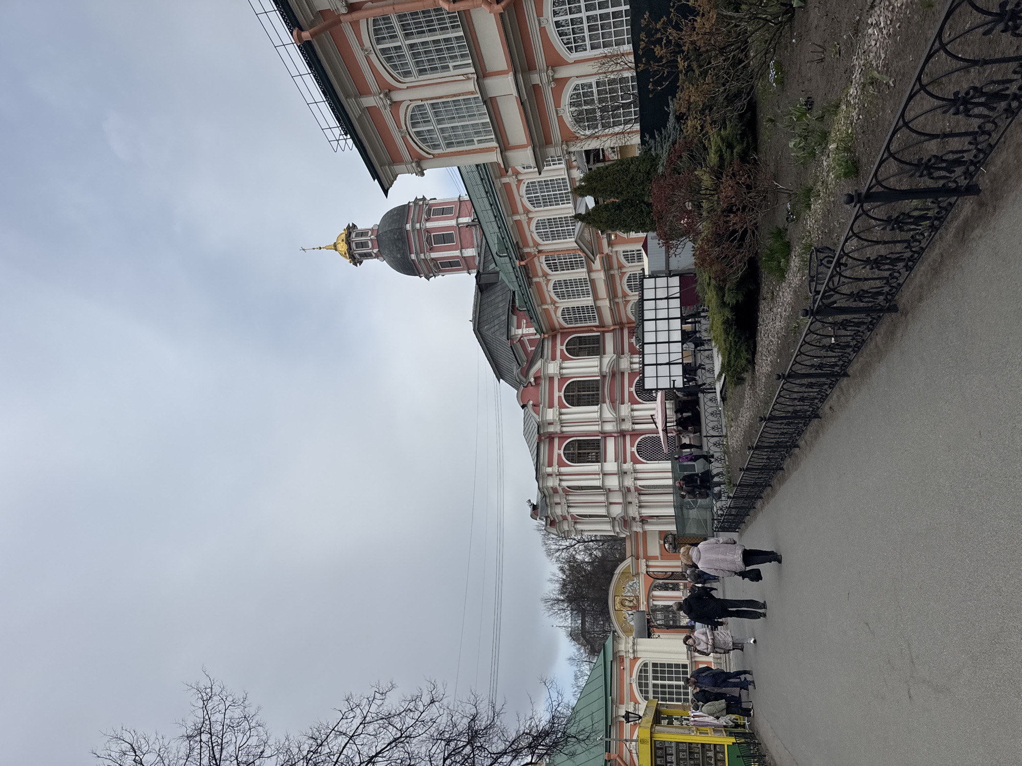 The image depicts a street scene in Saint Petersburg, Russia, in front of a large, ornate building with red and white facades and a prominent golden dome. The building's architectural style is reminiscent of Russian Orthodox churches and monasteries.
A paved walkway runs alongside the building, bordered by a decorative black iron fence. Several people are walking along the path.
The sky is overcast, suggesting a cloudy day. Bare trees are visible in the foreground, indicating a season with little or no foliage.