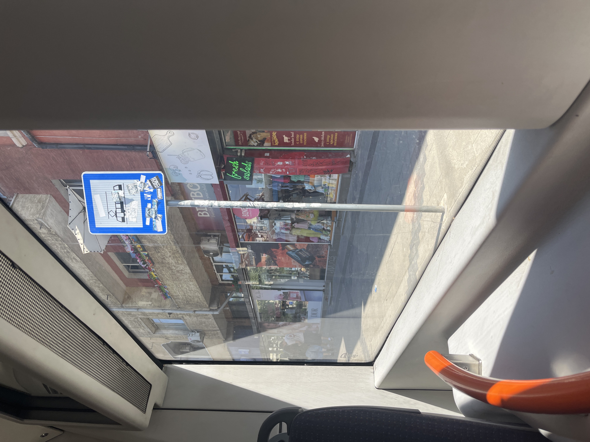 The image, taken from inside a public transport vehicle, shows a street scene in Sofia, Bulgaria. A blue traffic sign for a tram stop is visible, adorned with stickers. Beyond the tram stop, the street features shops with displays of clothing and food, including a sign that reads "fresh salads." Several people are walking on the sidewalk. The sunlight casts long shadows across the street, indicating it is likely midday. The interior of the transport vehicle, with a gray seat and an orange handrail, is also partially visible.