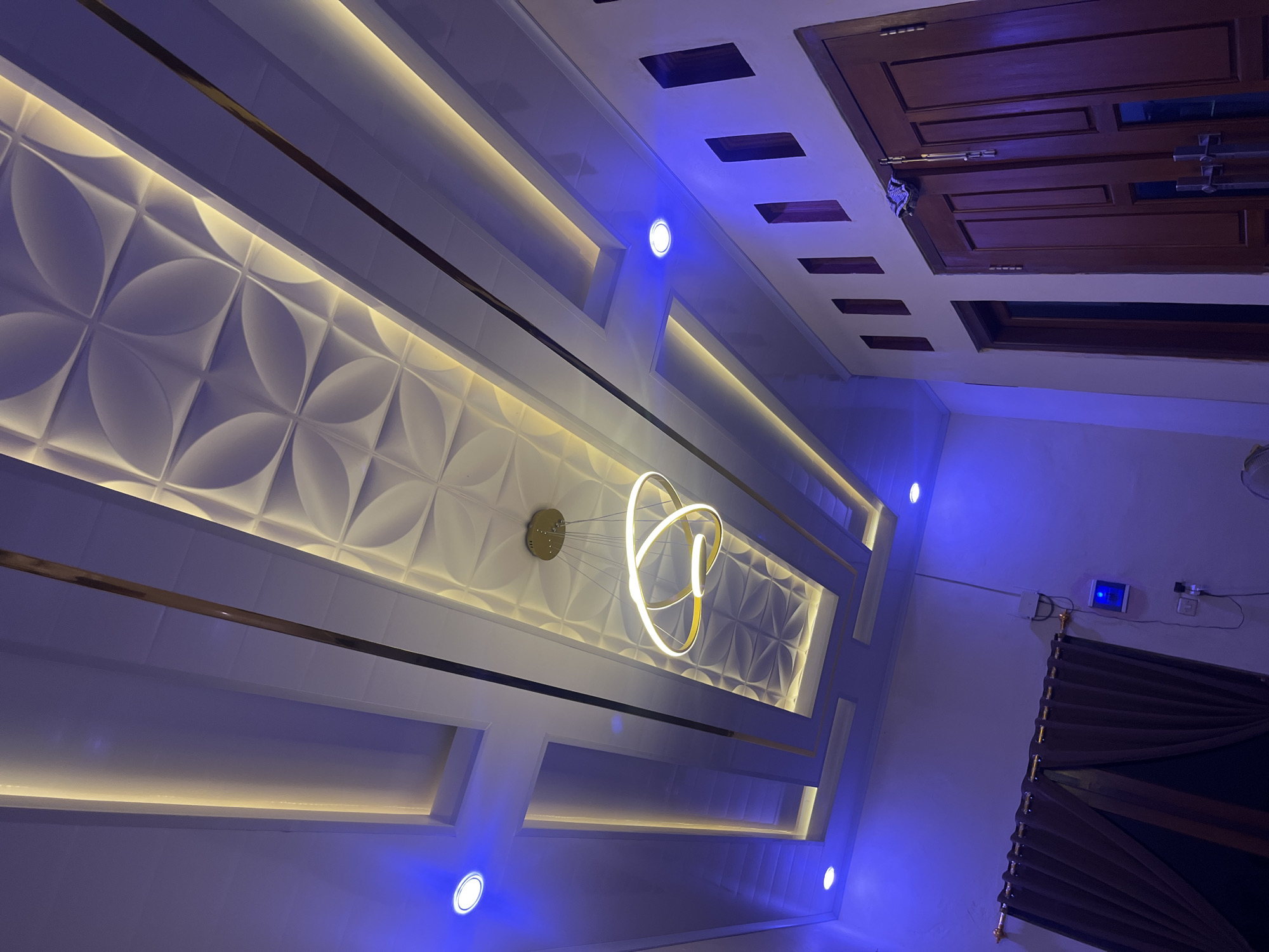 The image shows an interior view of a room with a modern ceiling design. The ceiling features recessed lighting with warm yellow light strips running along its edges and a central, textured panel with a floral pattern. A contemporary chandelier with gold rings hangs from the center of the ceiling. Blue LED spotlights are integrated into the ceiling, casting a cool glow on the room. On the right side of the image, a wall with several rectangular openings is visible, leading to a wooden double door. On the left side, dark brown curtains are drawn across a window. The room appears to be dimly lit, with the artificial lights being the primary source of illumination. The scene is likely within a residential or commercial building in Jatisari, Indonesia.