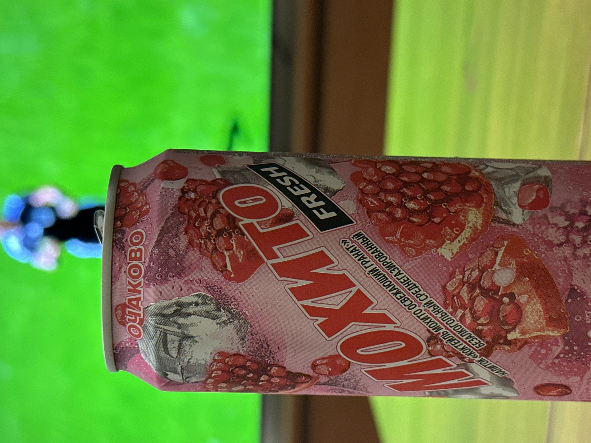 A close-up, eye-level shot shows a pink canned beverage with a label featuring pomegranate slices and the text "ОЧАКОВО" and "МОХИТО FRESH". The can is positioned in the foreground, resting on a light-colored, wooden surface. In the background, a blurred bright green area, likely a television screen, displays a shadowy figure. The overall scene is indoors with artificial lighting. The can's design suggests it is a drink from the Ochakovo company, potentially a non-alcoholic mojito flavored beverage. There is no identifiable information that places the scene specifically in Moscow, Russia.