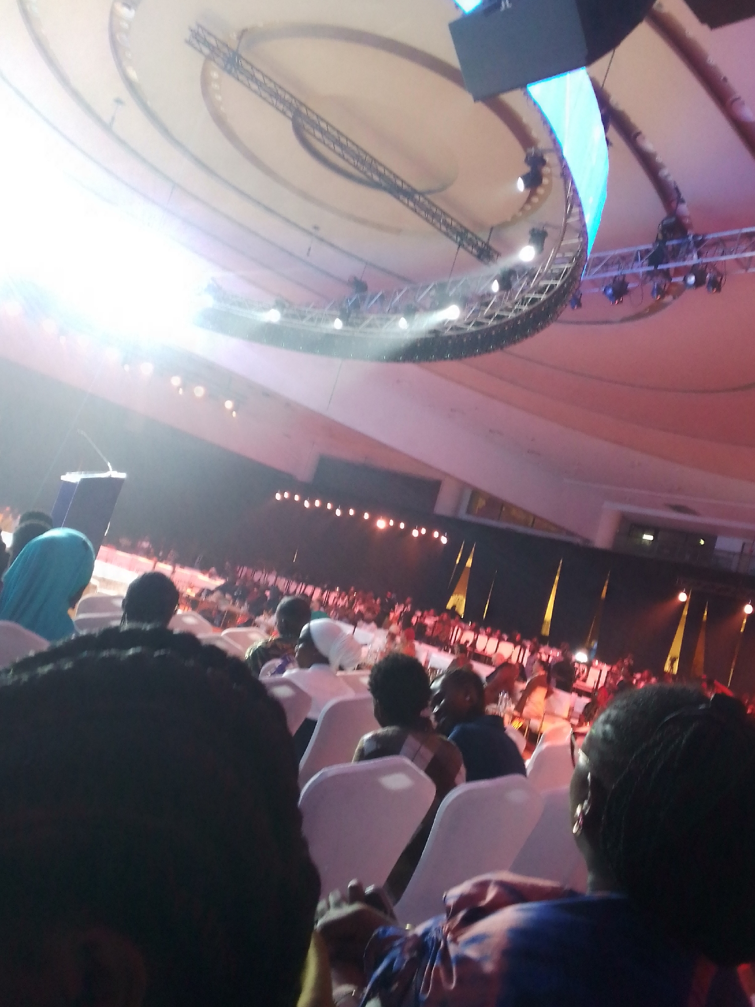 The image shows a large indoor gathering, likely a conference or event. Numerous people are seated in rows of white chairs, facing a stage area illuminated by bright lights. A lectern is visible on the left side of the frame, indicating a speaking engagement. Overhead, a circular lighting rig with multiple spotlights and a curved blue light display is suspended from the ceiling. The ceiling itself is a large, dome-like structure with visible architectural elements. The overall color scheme is warm, with red and orange hues from the stage lighting. The image is taken from the perspective of someone in the audience, with the heads of the people in front of them partially obscuring the view.