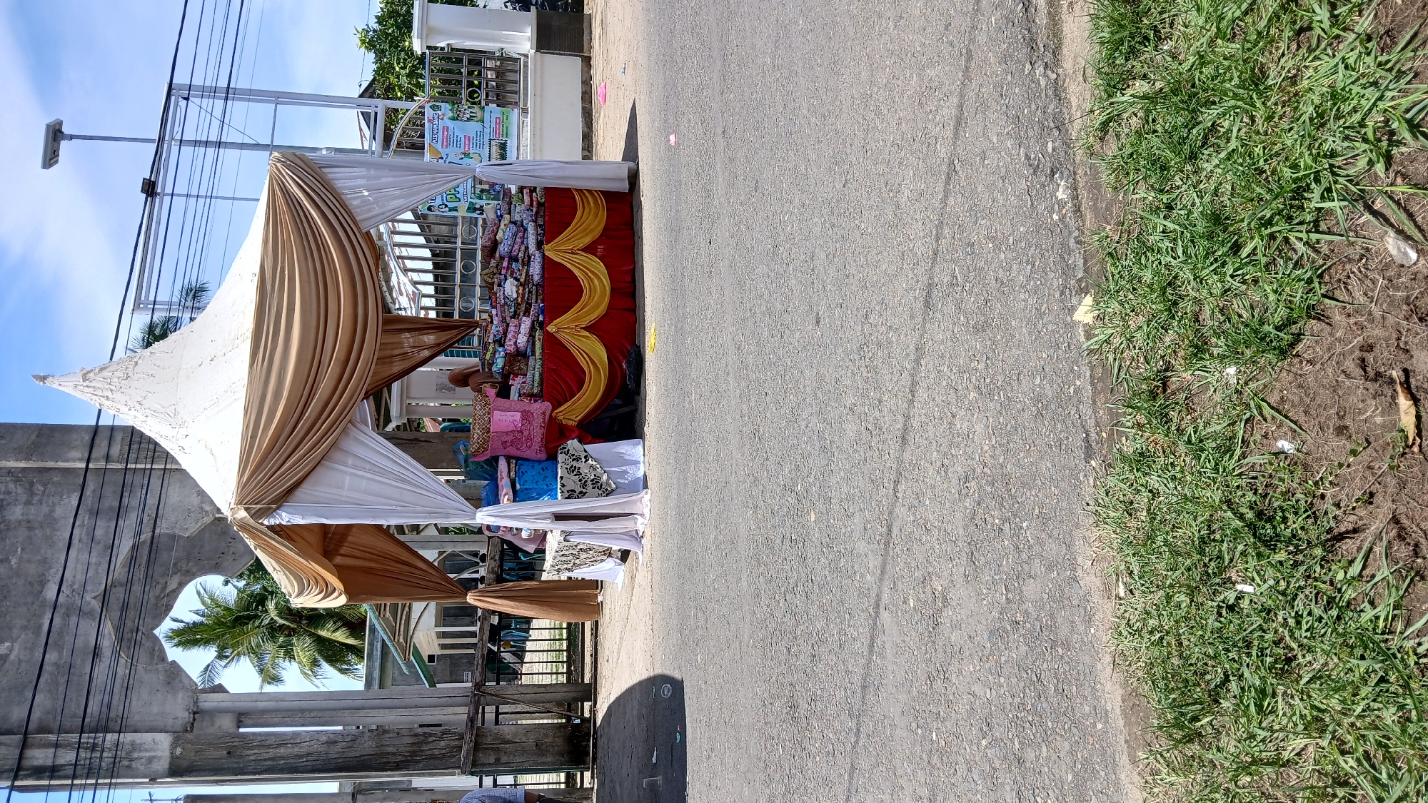 The image depicts a roadside vendor stall, likely in Lhokseumawe, Indonesia, given the cultural context. The stall is erected under a makeshift white and gold draped canopy. Merchandise, including cushions and other goods, is displayed. A red and gold skirt covers the table. The stall sits directly on the asphalt road, with a strip of grass bordering the edge of the pavement. Buildings and infrastructure are visible in the background, including a concrete archway and utility poles. The sky is visible and clear.