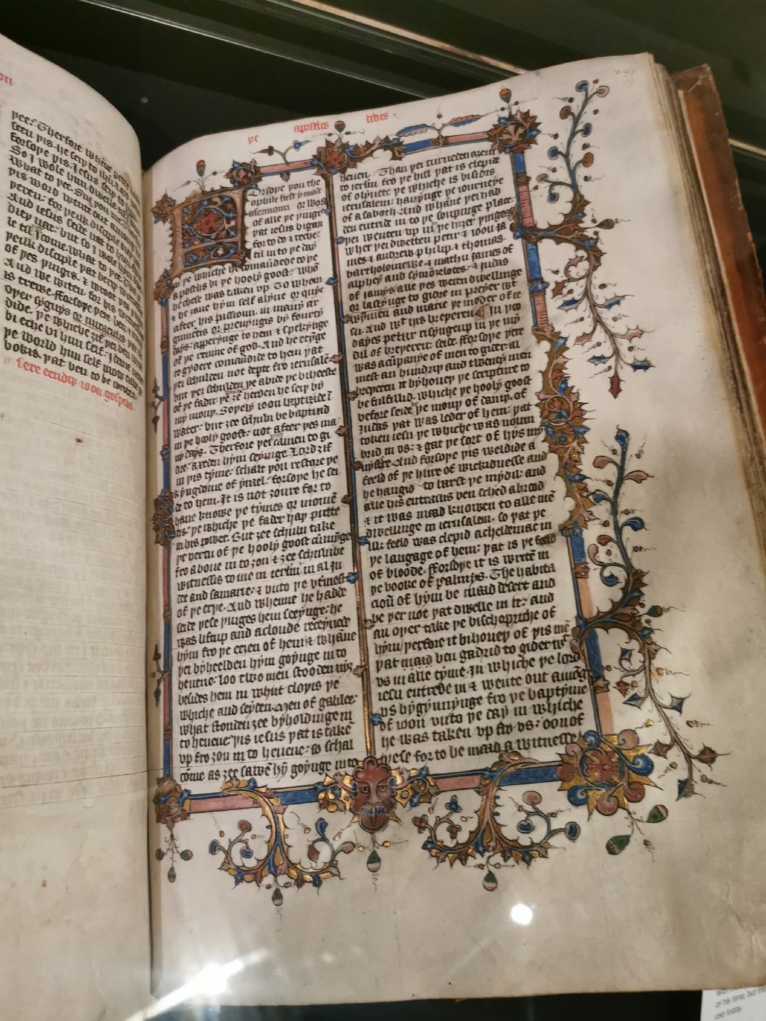 An open illuminated manuscript is displayed, likely in a library or museum setting against a dark background. The book is open to a two-page spread, revealing columns of text written in a Gothic or Blackletter script, primarily in dark ink. The language appears to be Middle English, characterized by words like "pe" (the), "youre" (your), and distinct letterforms.
The pages are richly decorated with intricate illumination. The left page features a large, decorated initial letter, possibly a 'T' or 'P', and a gold-leaf border extending down the inner margin. The right page also has a large decorated initial, likely an 'H', accompanied by an ornate border that frames the outer and bottom margins of the text block. These borders are elaborate, featuring swirling vine-scroll designs, acanthus leaves, small roundels, and what appear to be grotesque human or animal faces integrated into the foliage. Gold leaf is extensively used throughout the illuminations, catching the light and adding a shimmering effect, complemented by blue, red, and green pigments.
The text is divided into columns, and the top right corner of the right page displays the page number "297". Headings such as "agonies" and "tubes" are visible above the text on the right page, suggesting specific sections or chapters. The manuscript is in good condition for its age, showcasing detailed craftsmanship and preservation.