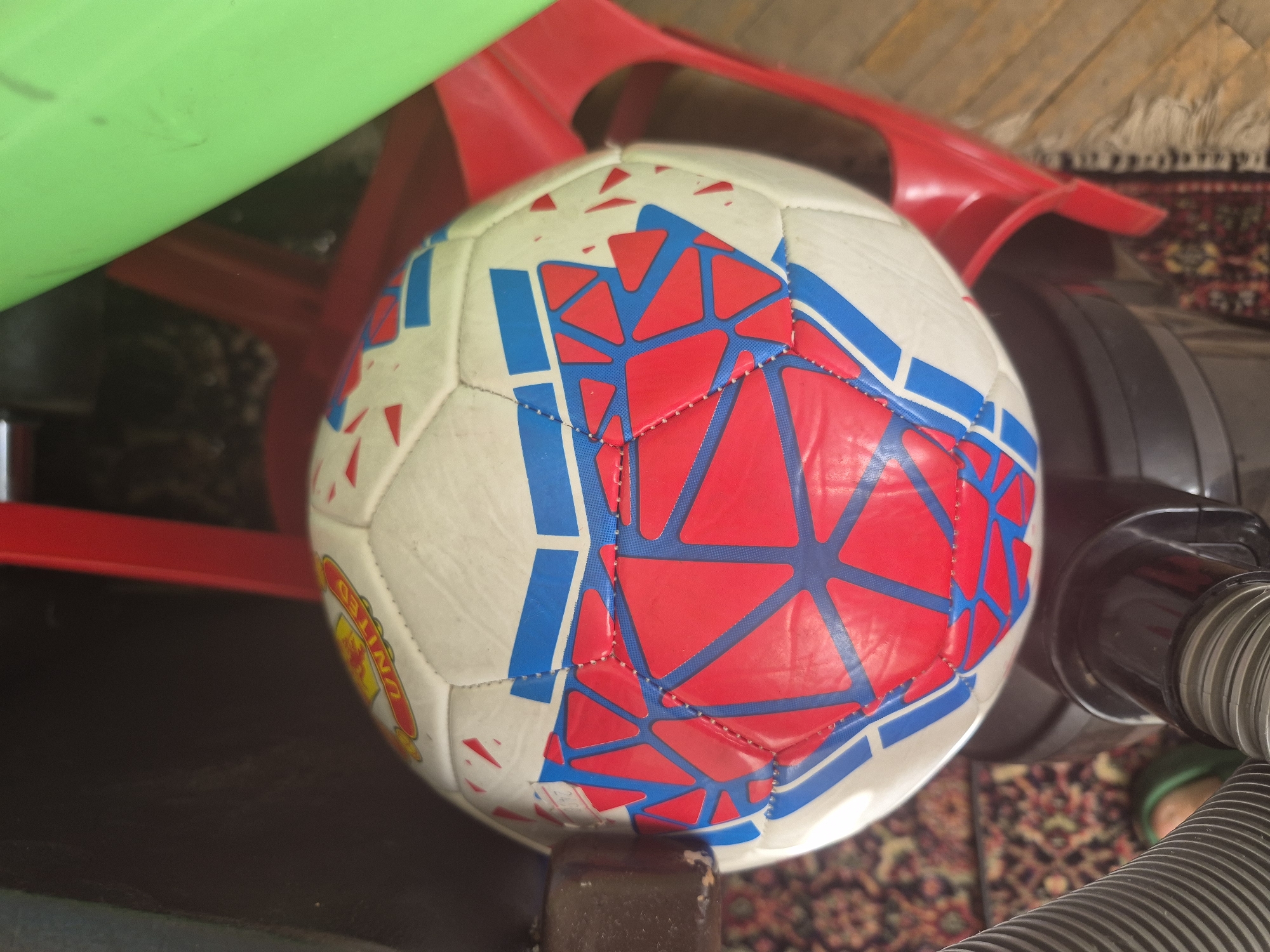 A close-up, eye-level shot captures a soccer ball positioned indoors, likely in a home setting in Yerevan, Armenia. The ball, which is the central focus, is predominantly white with striking red and blue geometric patterns. The red areas feature an intricate web of darker red lines, creating a textured or segmented appearance. The blue sections are solid.
To the left side of the ball, a bright green plastic object, possibly part of a toy or furniture, is visible, partially obscuring a red plastic object behind it. Another red plastic item, possibly a chair leg or frame, is seen extending from the middle-left towards the upper-right background. On the right side of the ball, a dark grey or black object, resembling a vacuum cleaner's hose and body, is partially visible, extending towards the bottom right corner. The soccer ball itself rests on a dark brown, possibly wooden, object that appears to be a furniture leg or support.
A patterned rug or carpet with muted tones of red, cream, and brown covers the floor in the background, providing a domestic context. On the left side of the ball, a yellow and red logo is partially visible, featuring the word "UNITED," strongly suggesting a Manchester United brand ball. Another small, illegible white text or number is barely discernible on the lower-left white panel of the ball. The lighting appears natural and even, suggesting daytime. There is no discernible activity taking place; the scene is static.