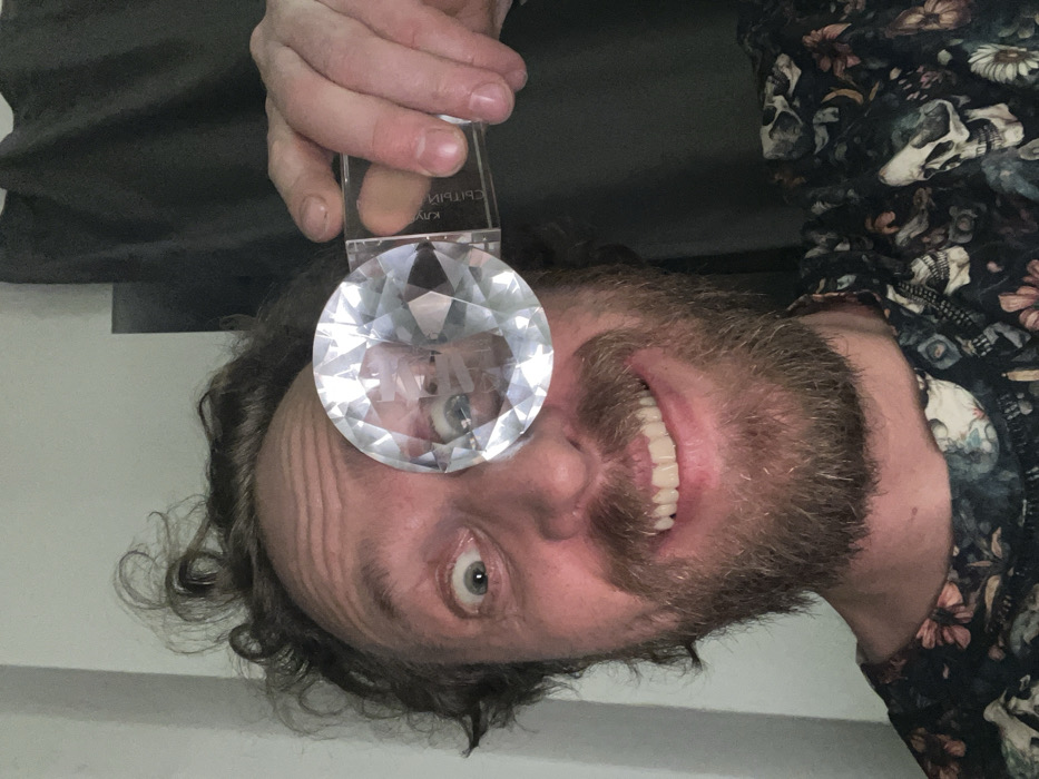 A man with curly hair and a beard smiles, teeth showing. He is holding a large, clear, faceted gem directly in front of one eye, distorting his iris in the reflection. He wears a dark floral and skull-patterned shirt. A hand holding the gem’s plastic container is visible above it. The background includes a grey wall and what appears to be dark green fabric or clothing.