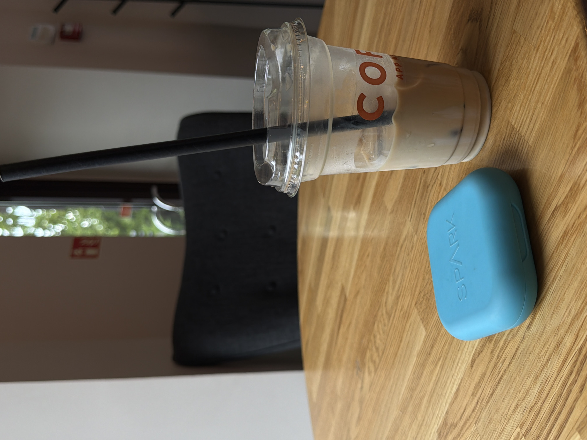 The image shows a Costa Coffee cup with a black straw and a blue, rectangular object with "SPARK" written on it, both sitting on a wooden table. The background includes a chair and a window with a blurred outdoor view, suggesting an indoor setting. The location may be Riga, Latvia, but there is not sufficient information to confirm this from the image.