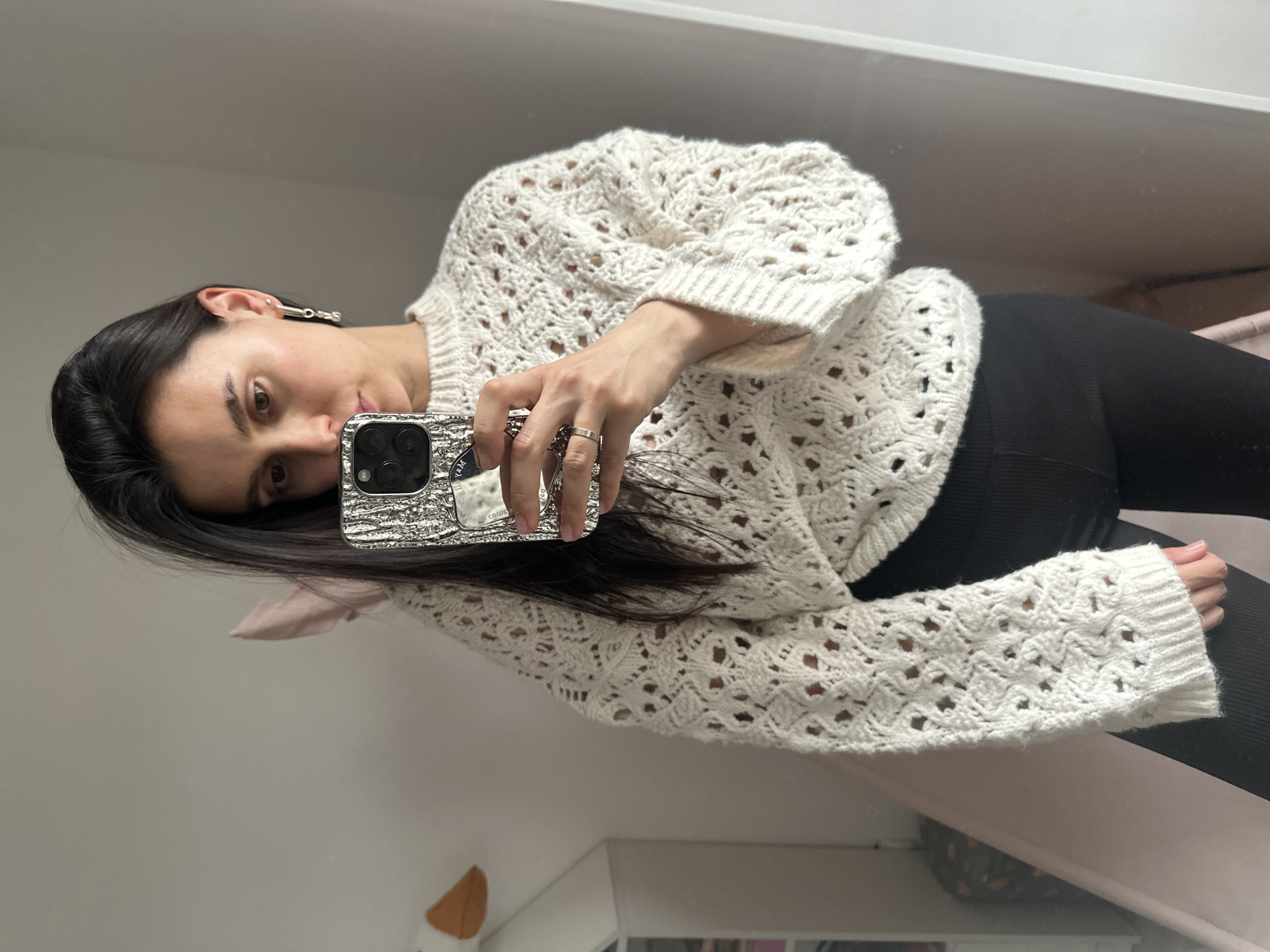 A woman with dark hair and a white sweater is taking a selfie in a mirror. She is wearing black leggings. The background appears to be a room with a bed and a white cabinet. The location is unknown and there is no information indicating it is Chaikovsky, Russia.